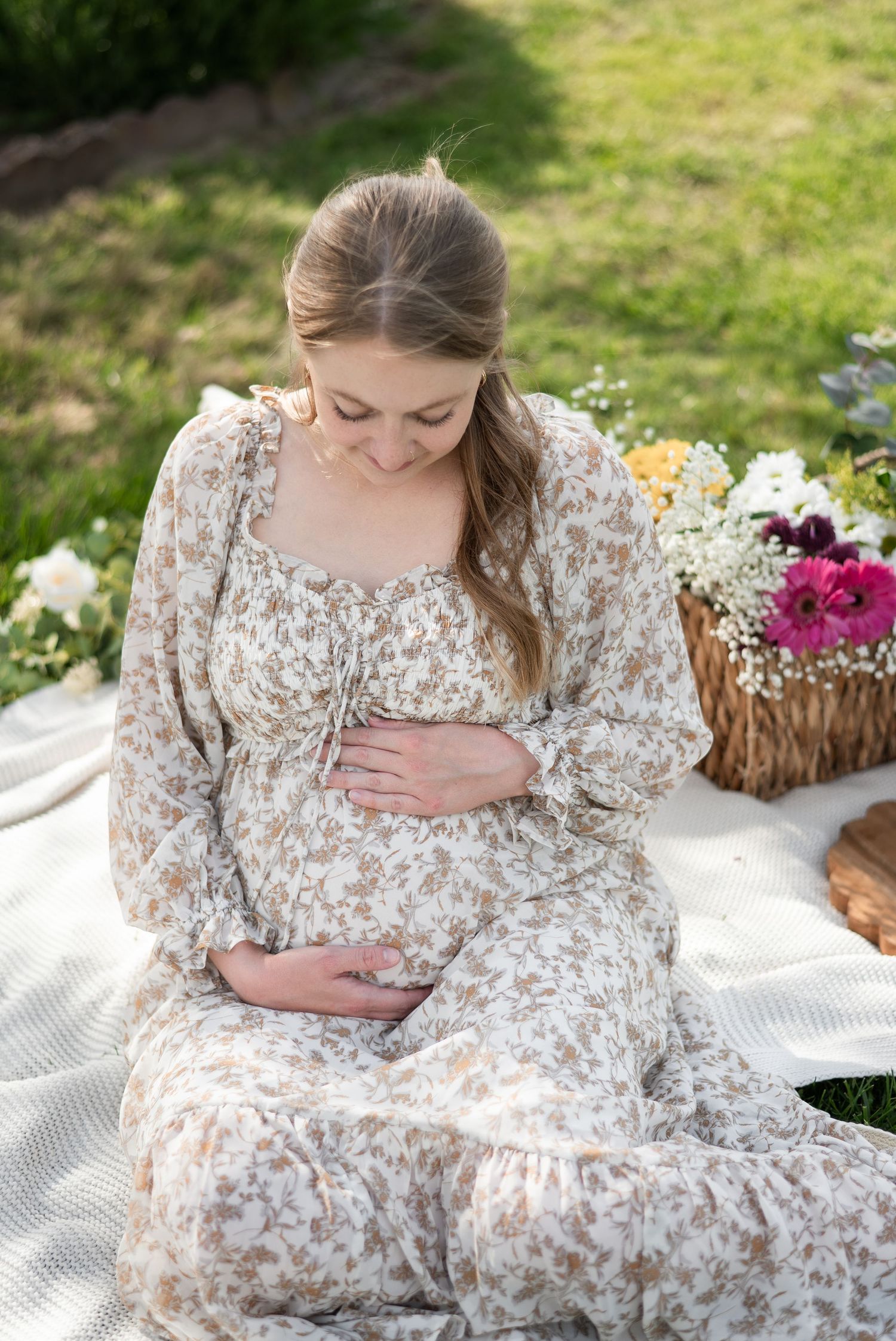Expecting mother in a floral dress sits on white blanket during outdoor maternity photoshoot with flower basket nearby.