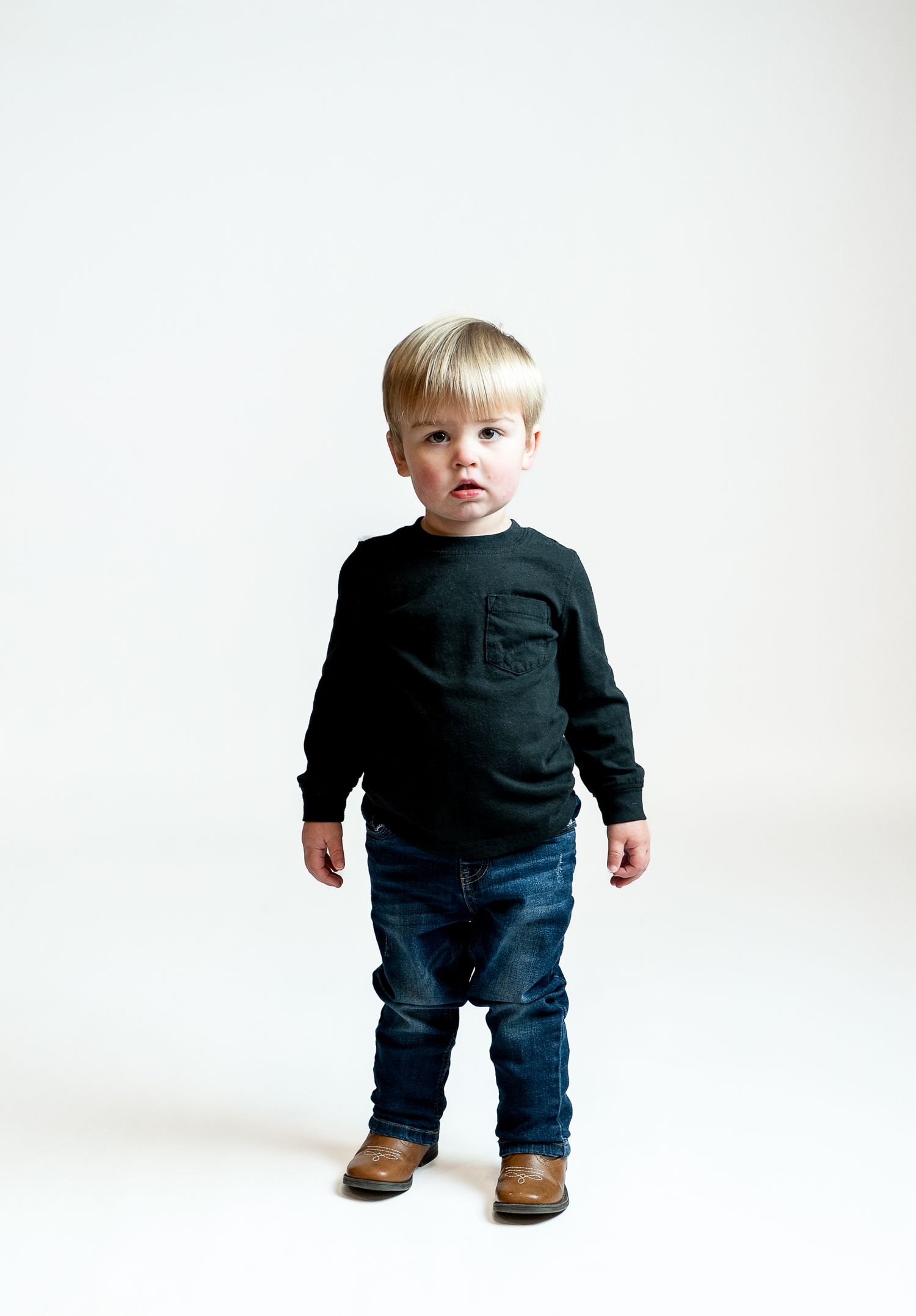 A toddler wearing a black sweater and jeans poses against a white studio background.