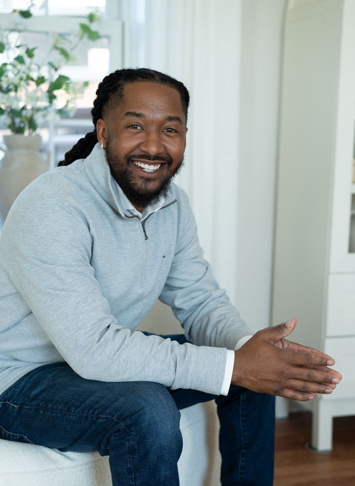 Smiling man with dreadlocks wearing a gray sweatshirt and jeans, sitting relaxed in a bright modern interior.
