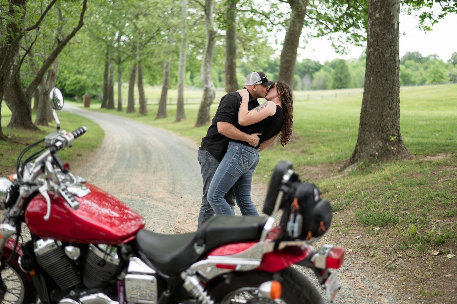 A couple embraces on a tree-lined path next to a parked red motorcycle in a scenic park setting.