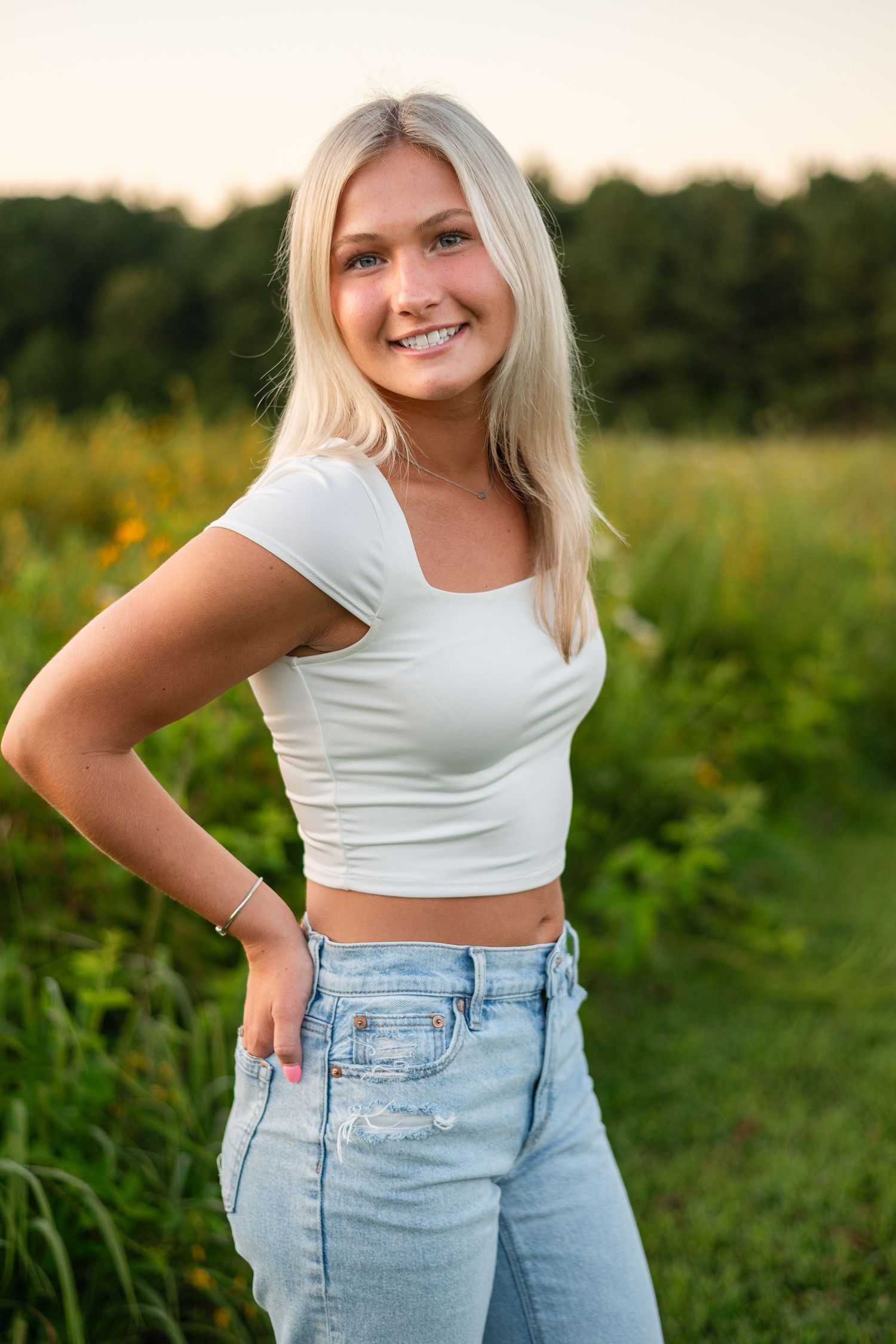 A person wearing a white crop top and light wash jeans poses outdoors in a green field during golden hour.