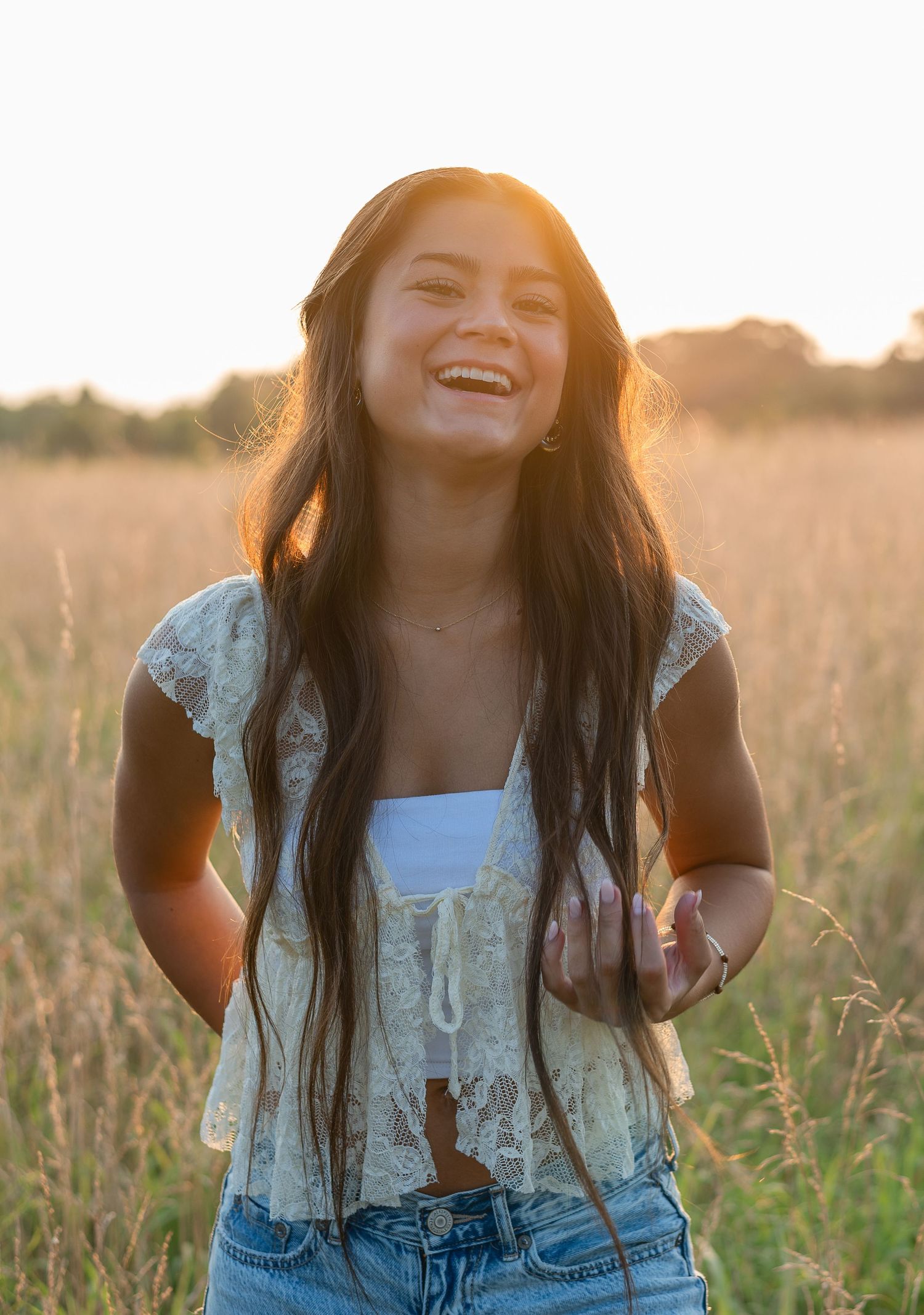 Young woman with long dark hair laughs joyfully in a golden sunlit field, wearing a lace top and denim shorts.