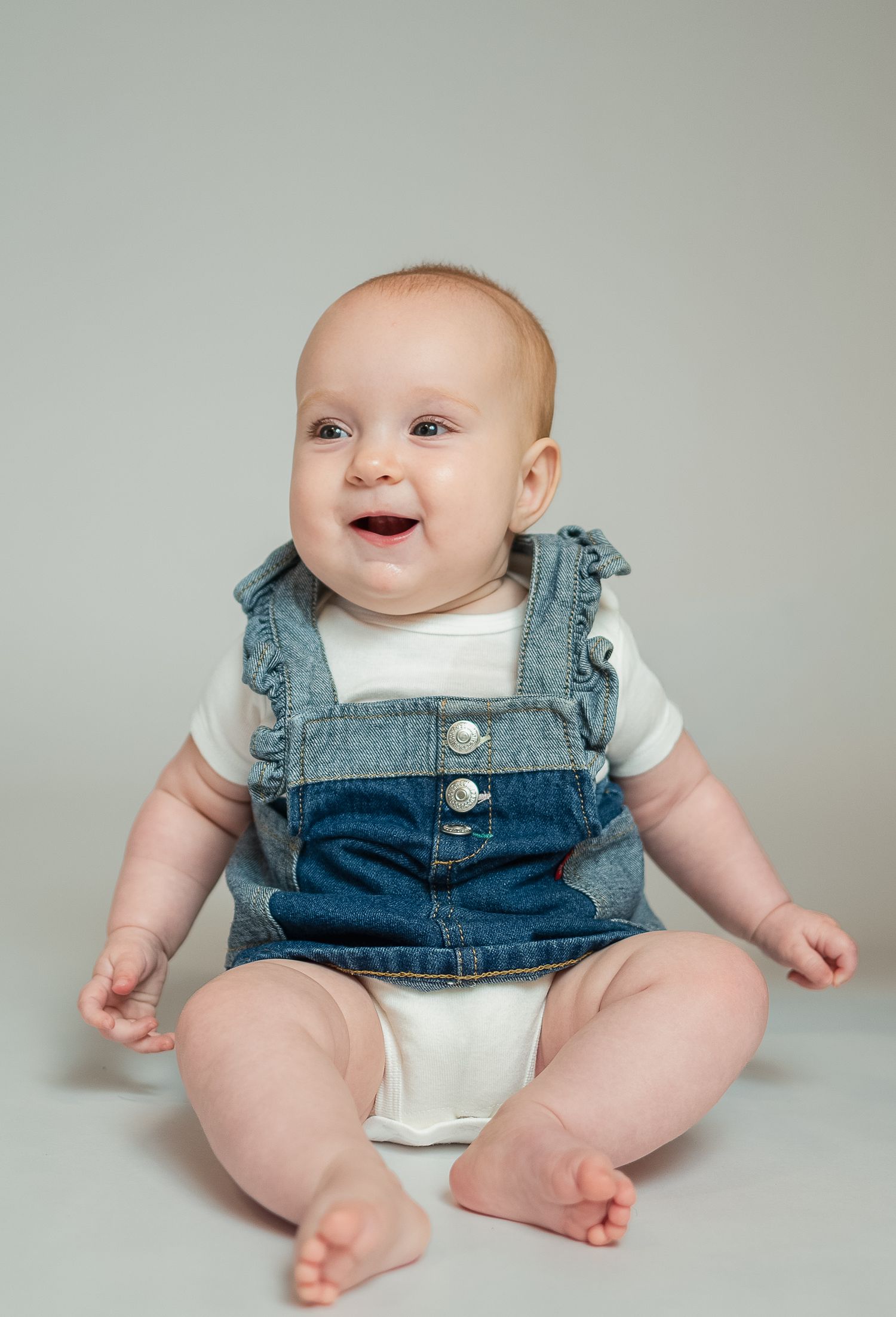 Smiling baby in denim overalls and white onesie sitting on a light gray background.