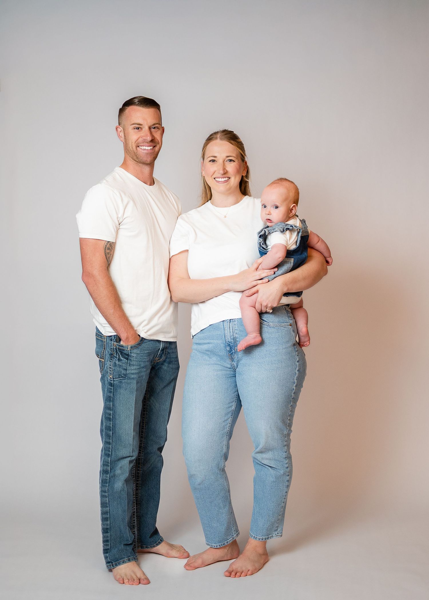 A young family in white tees and jeans poses barefoot in a studio, with the mother holding a baby.