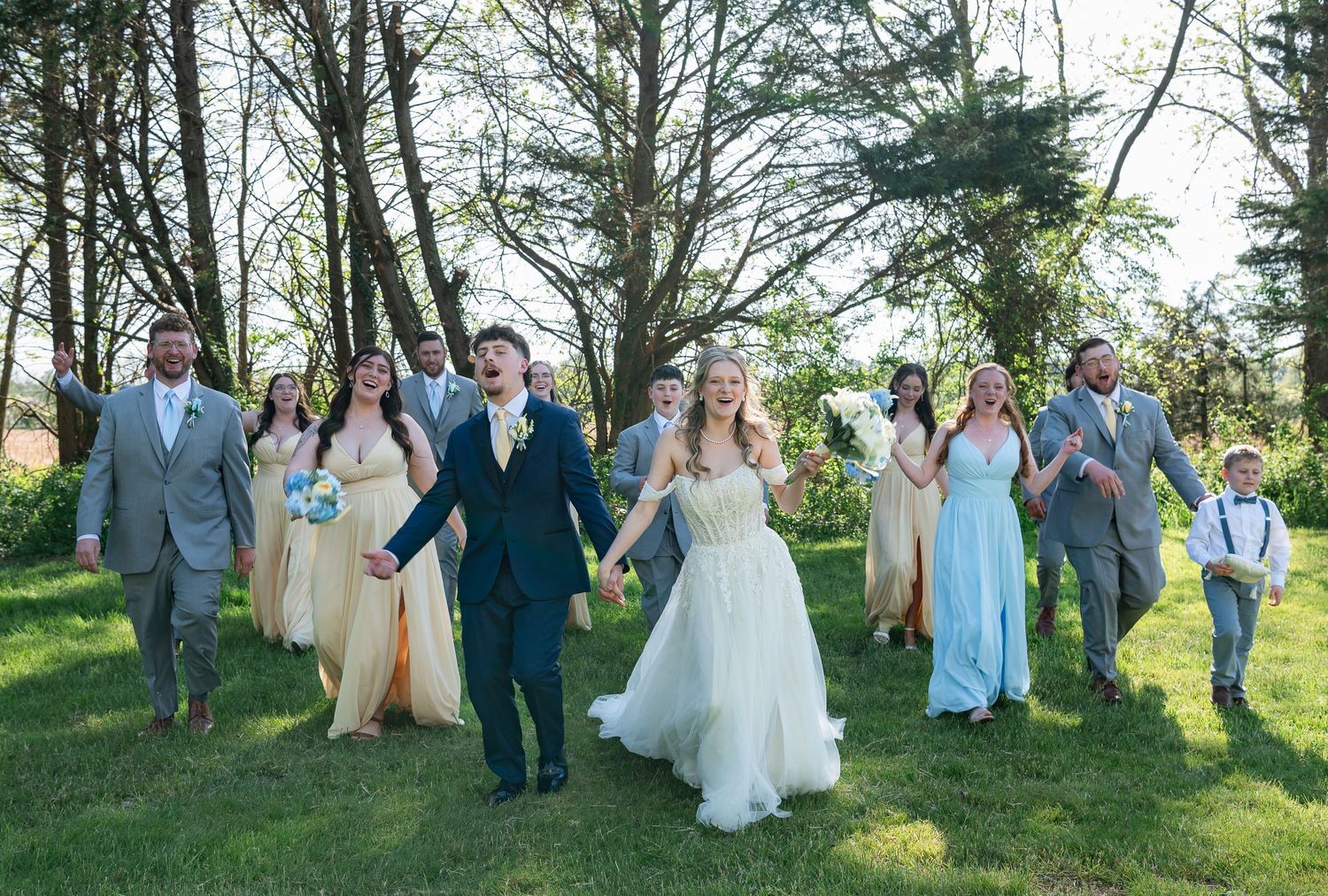Wedding party walking outdoors on green grass, bride in white gown, groomsmen in gray suits, bridesmaids in pastel dresses.