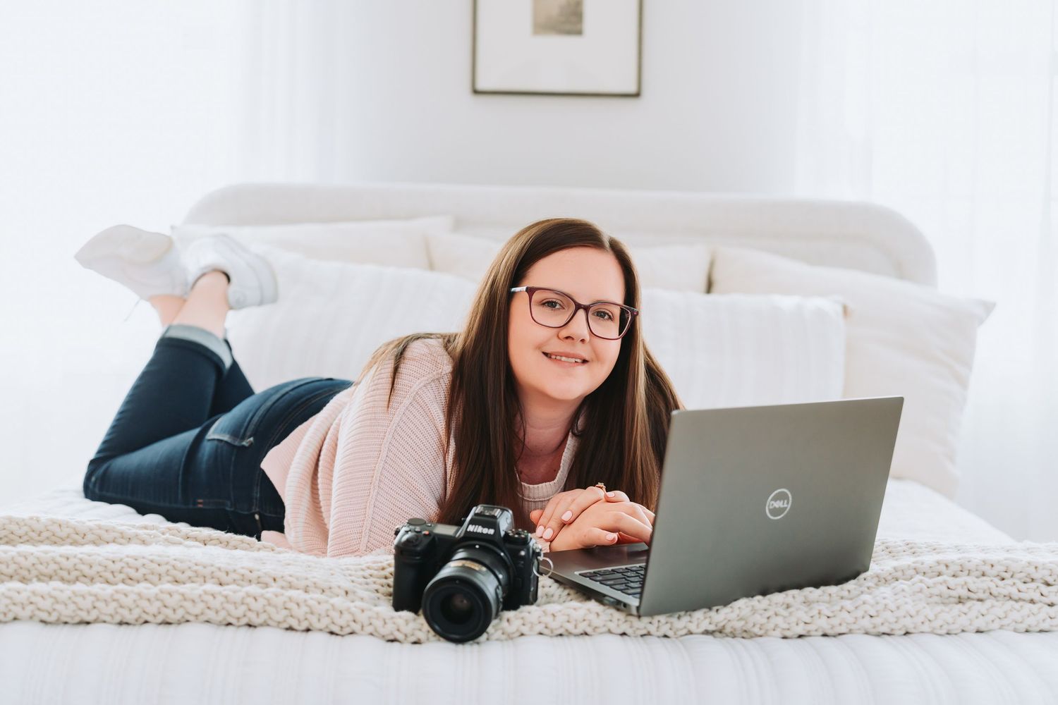 Someone lying on bed working on Dell laptop with camera nearby wearing pink sweater and glasses.