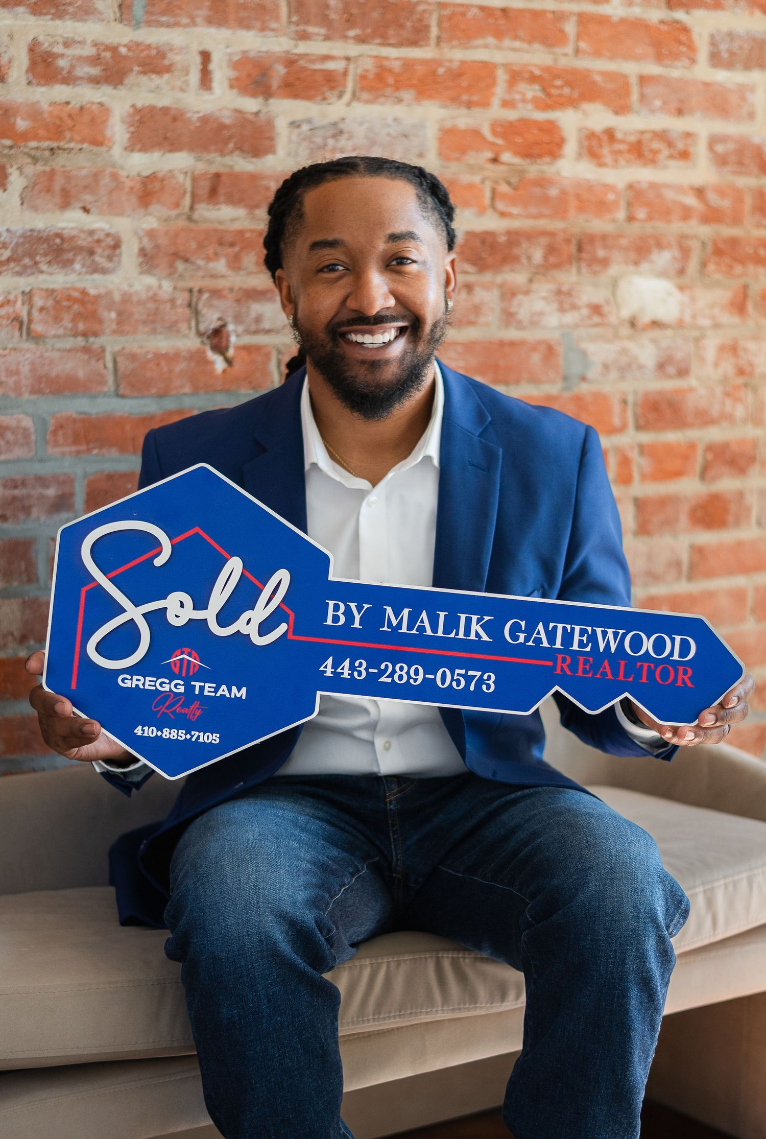 Malik Gatewood, Realtor from Gregg Team, smiling and holding a blue Sold sign, wearing a blue blazer.