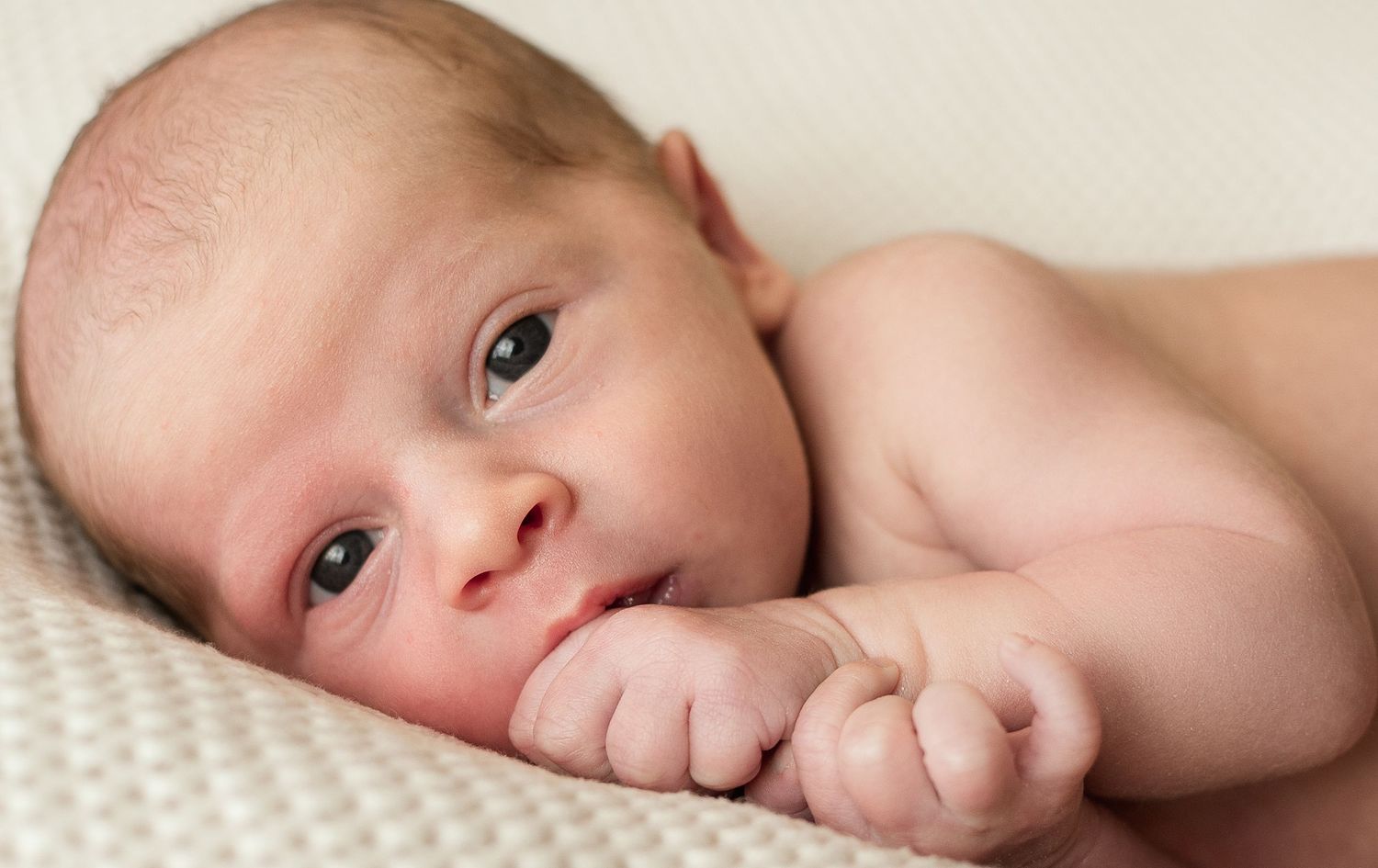 A newborn baby lies on a white blanket while sucking on their thumb.