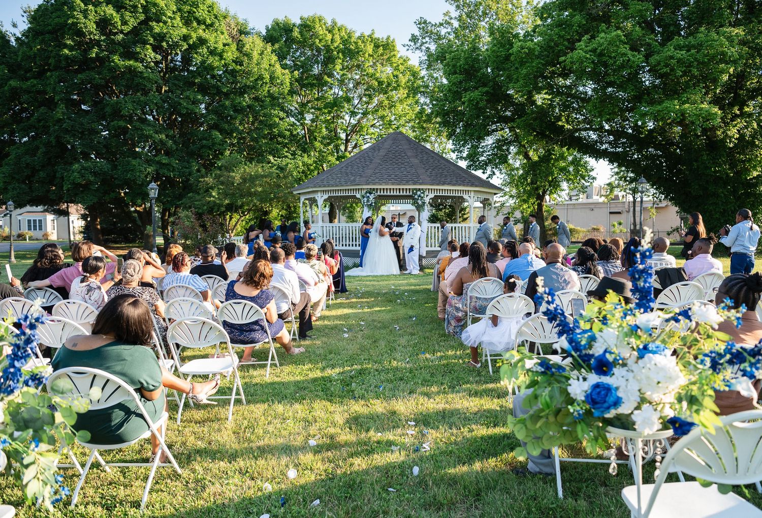 Outdoor wedding ceremony on green lawn with white chairs, blue floral arrangements, and a classic gazebo altar under clear blue skies.