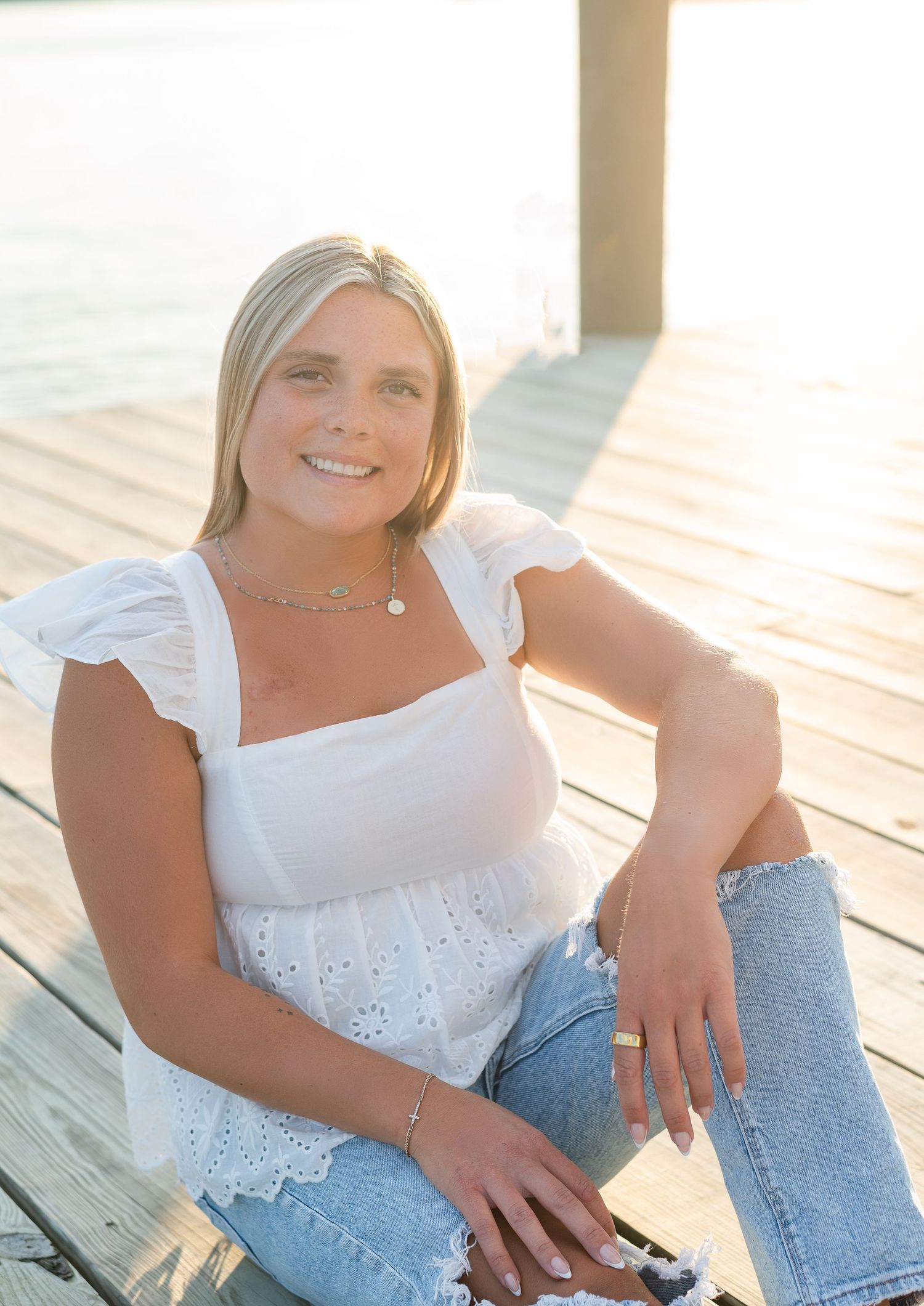 Smiling blonde woman in white eyelet top and jeans sitting on a sunlit wooden dock near water.