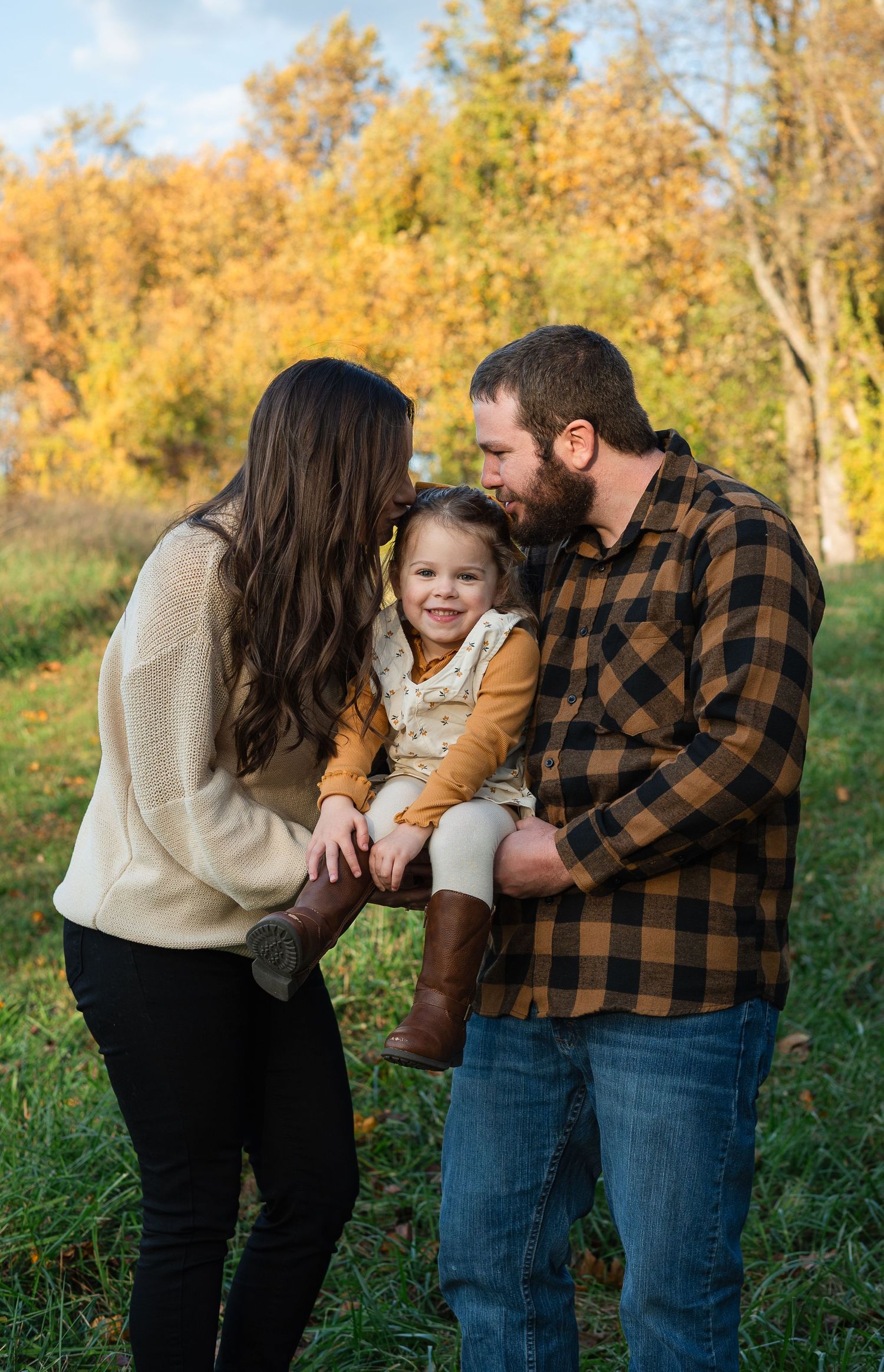 Family shares tender moment together in autumn field with golden trees in background.