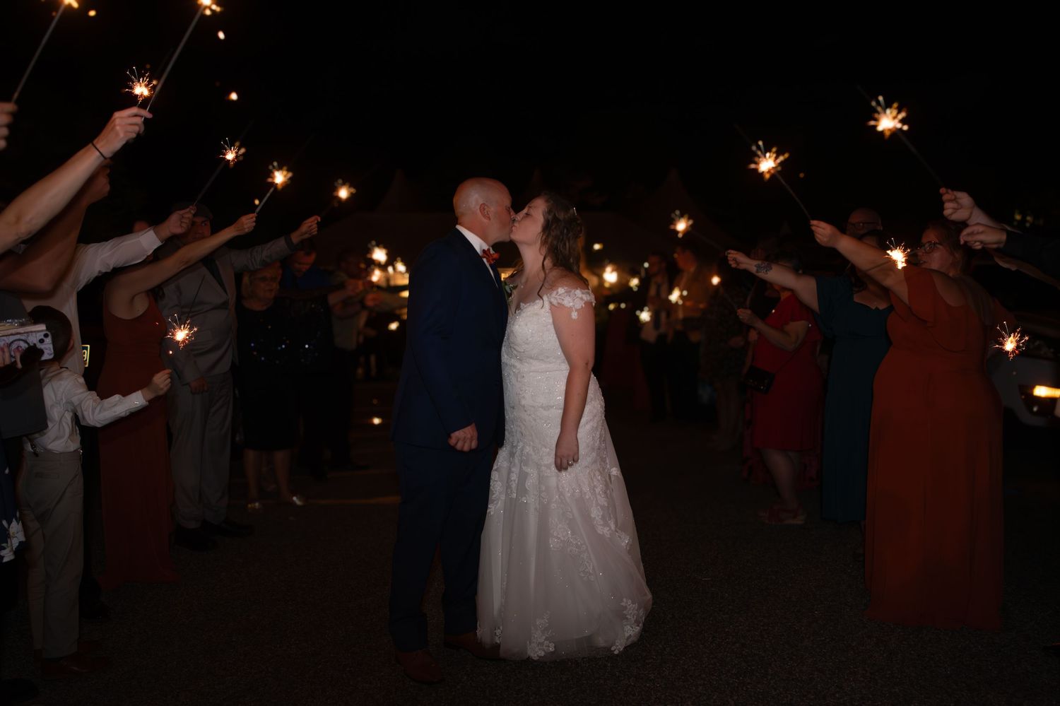 Wedding couple kiss during sparkler exit with guests holding lights at night.