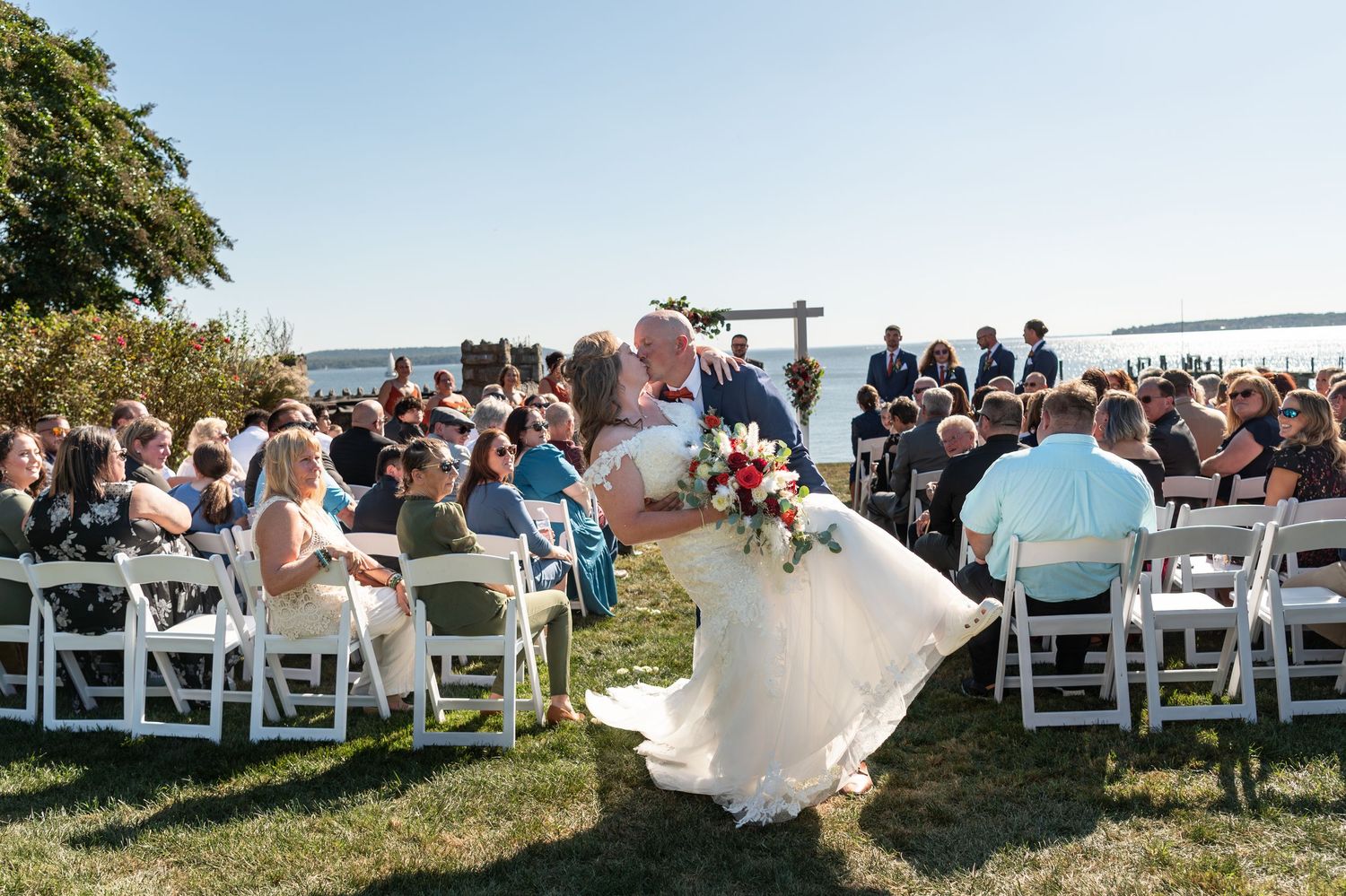 A scenic outdoor wedding ceremony takes place on a grassy waterfront area with guests seated in white folding chairs.