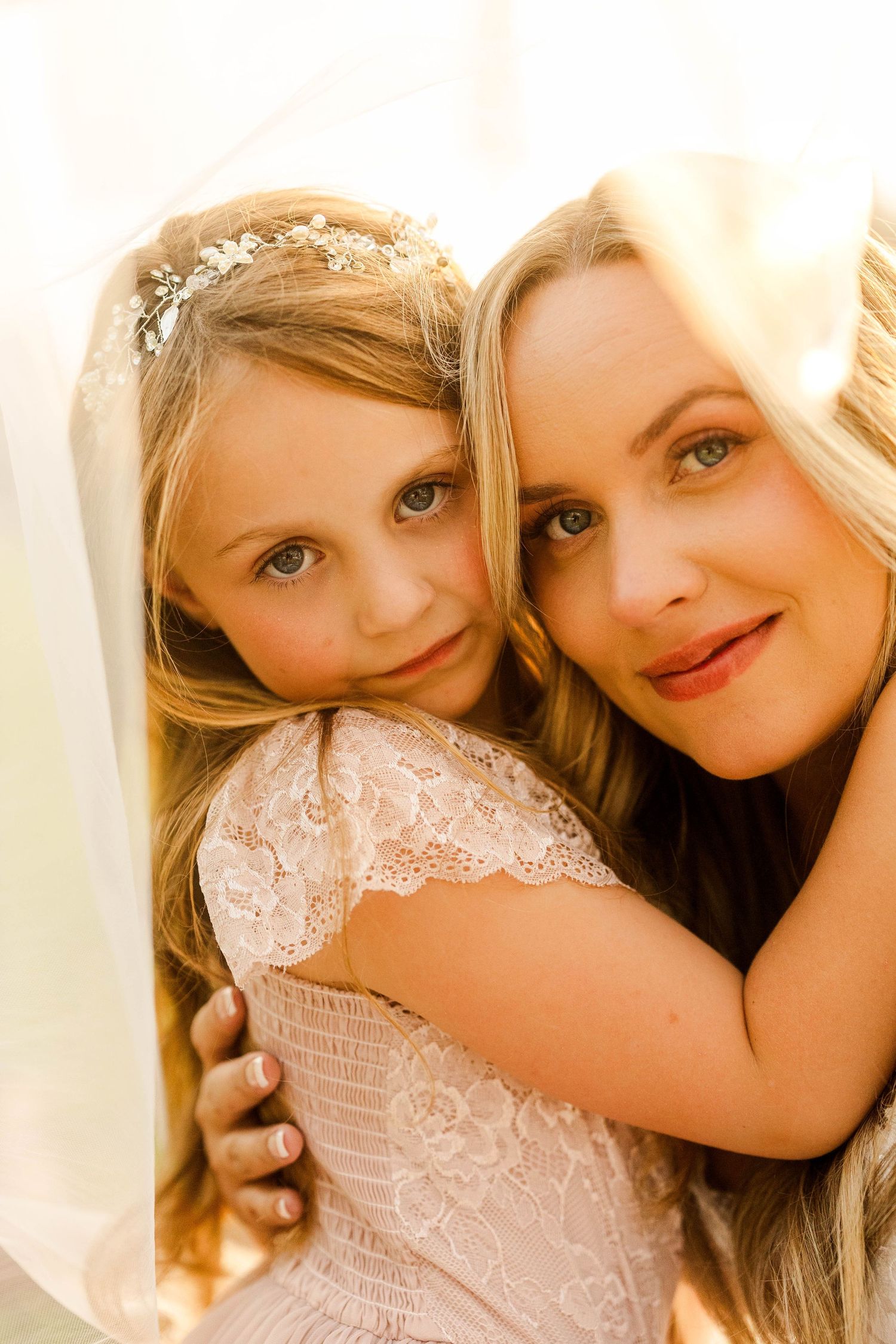 Close-up of bride and flower girl with floral crown smiling together under a white wedding veil.