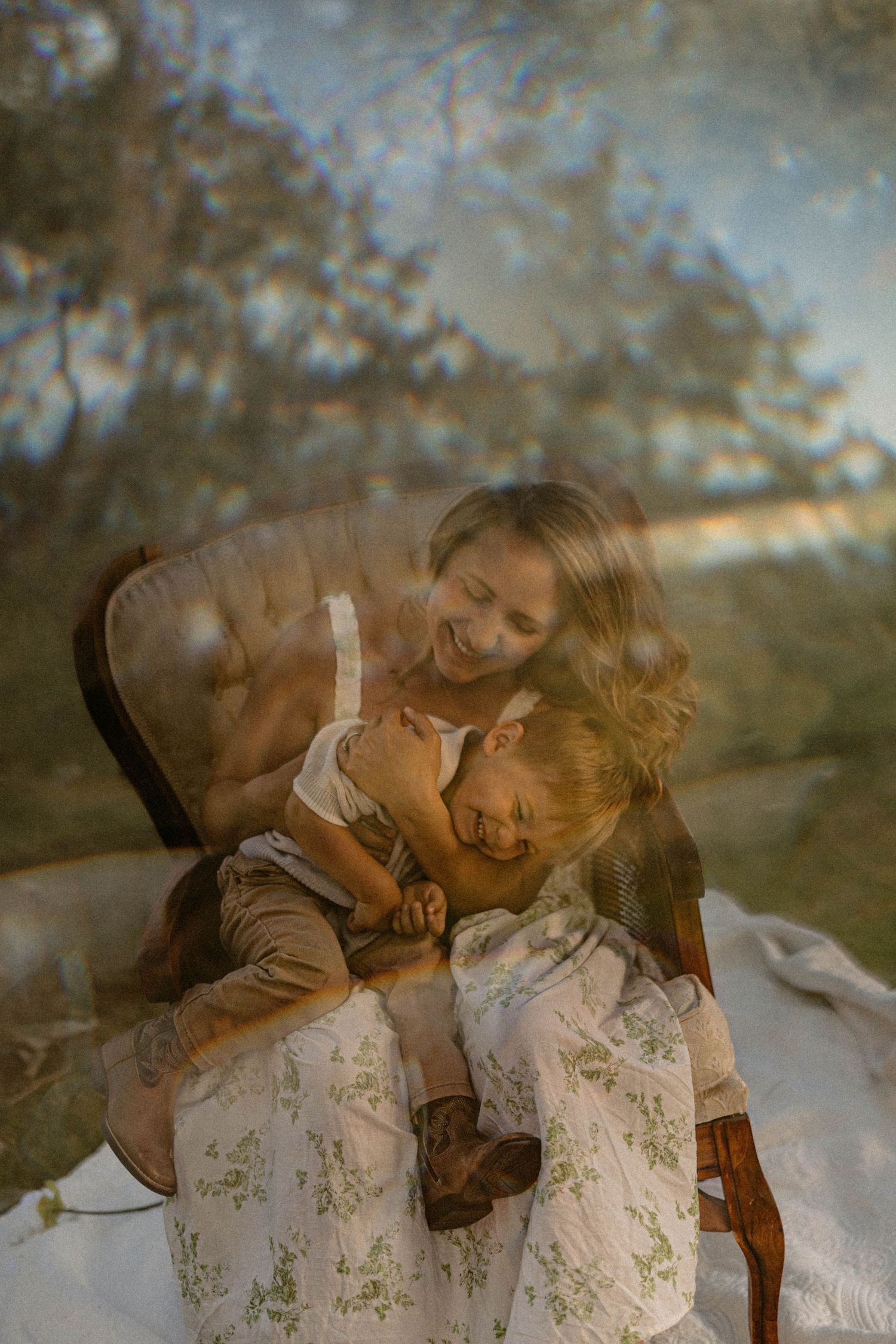 A joyful mother in a floral dress laughs with her toddler on her lap in a sun-drenched outdoor setting.