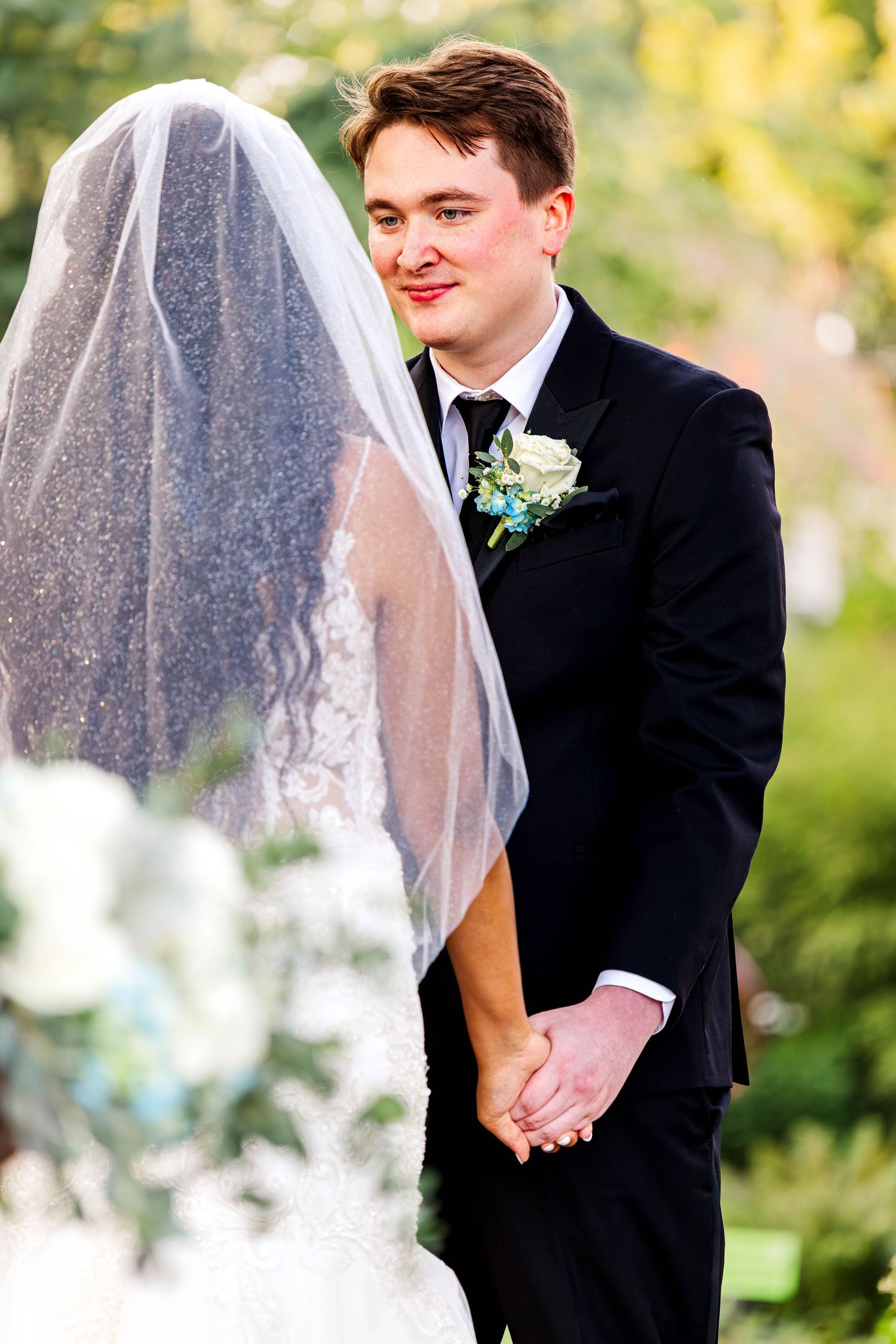 A groom in a black tuxedo holds his bride's hand as her veil flows in an outdoor garden ceremony.