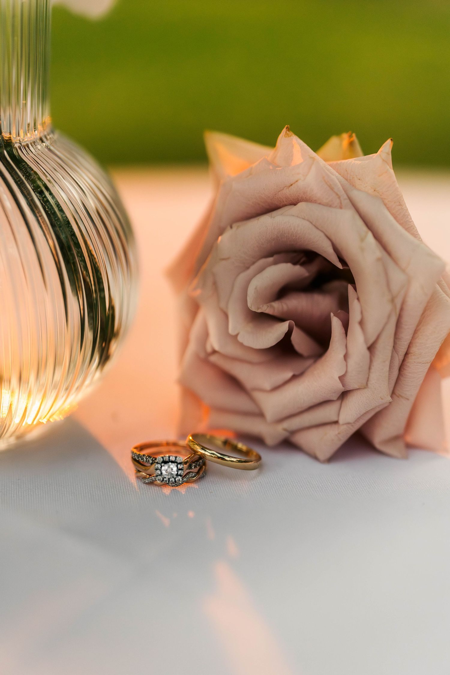 Gold wedding rings beside a blush pink rose and glass vase on a white reflective surface.