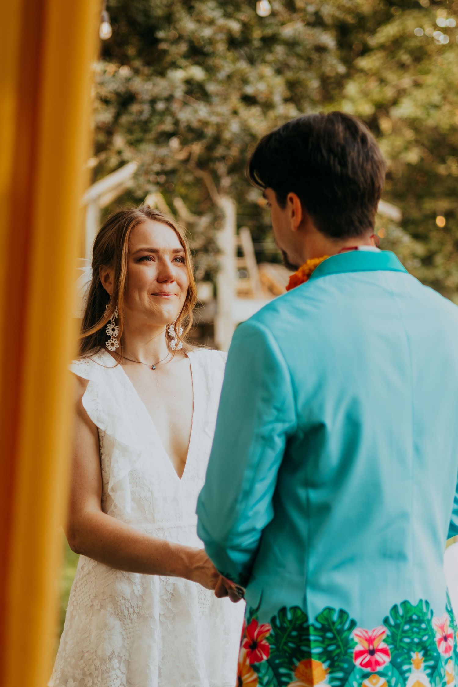 Tearful bride in a white lace dress exchanges vows with her groom in a colorful tropical wedding outfit.