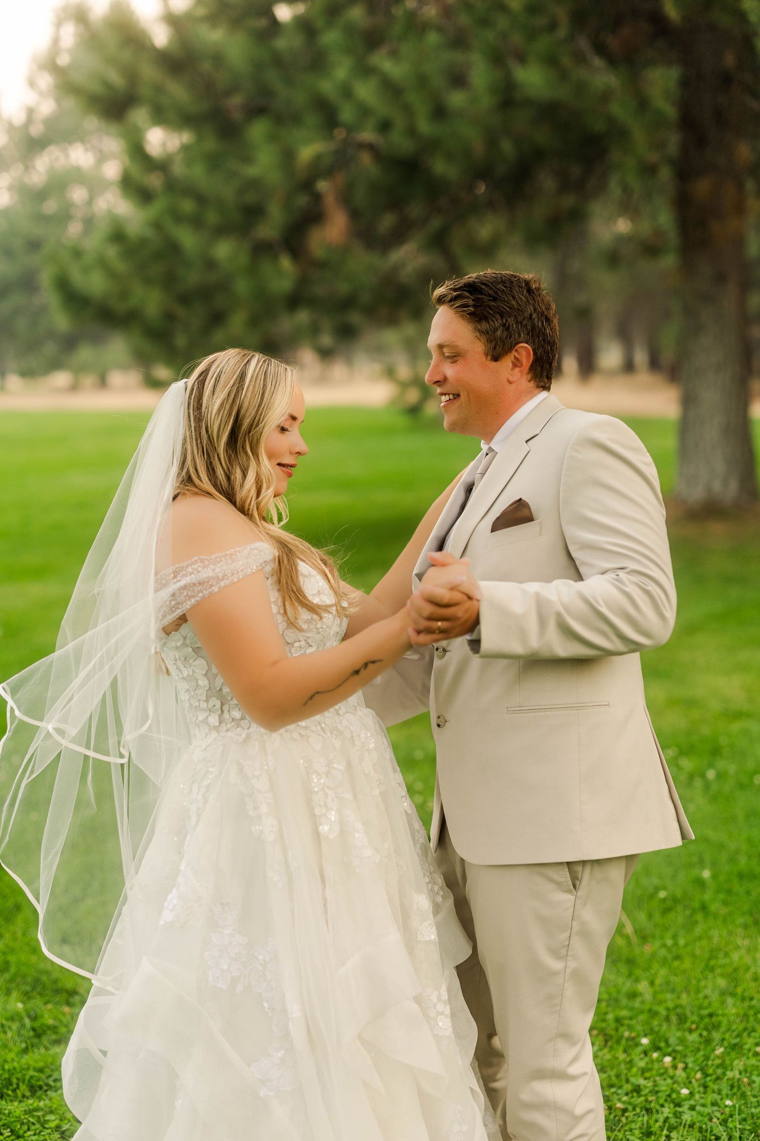 Smiling bride in off-shoulder gown and groom in tan suit sharing a joyful moment outdoors.