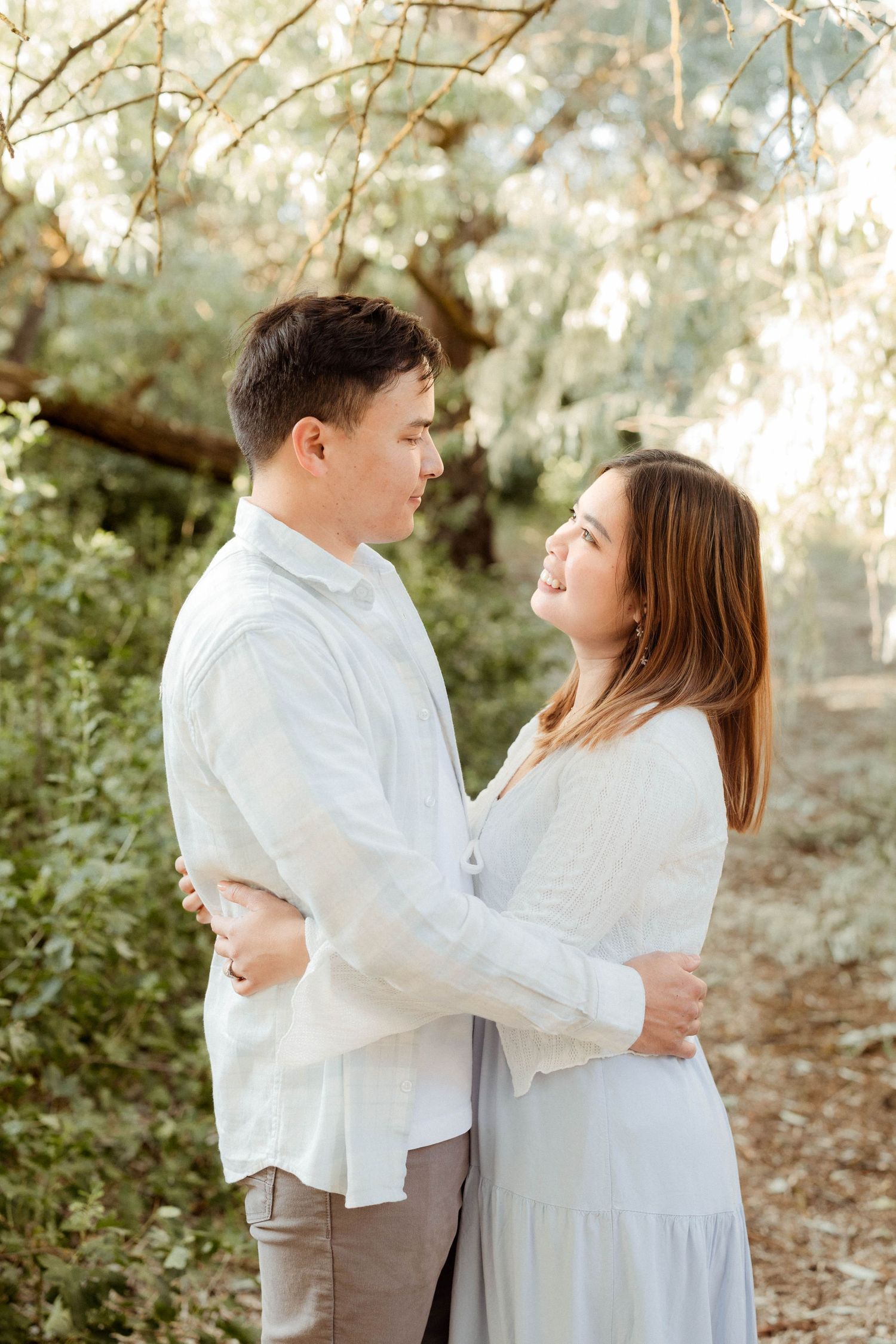 A young couple gazes at each other lovingly while embracing under sunlit trees outdoors.