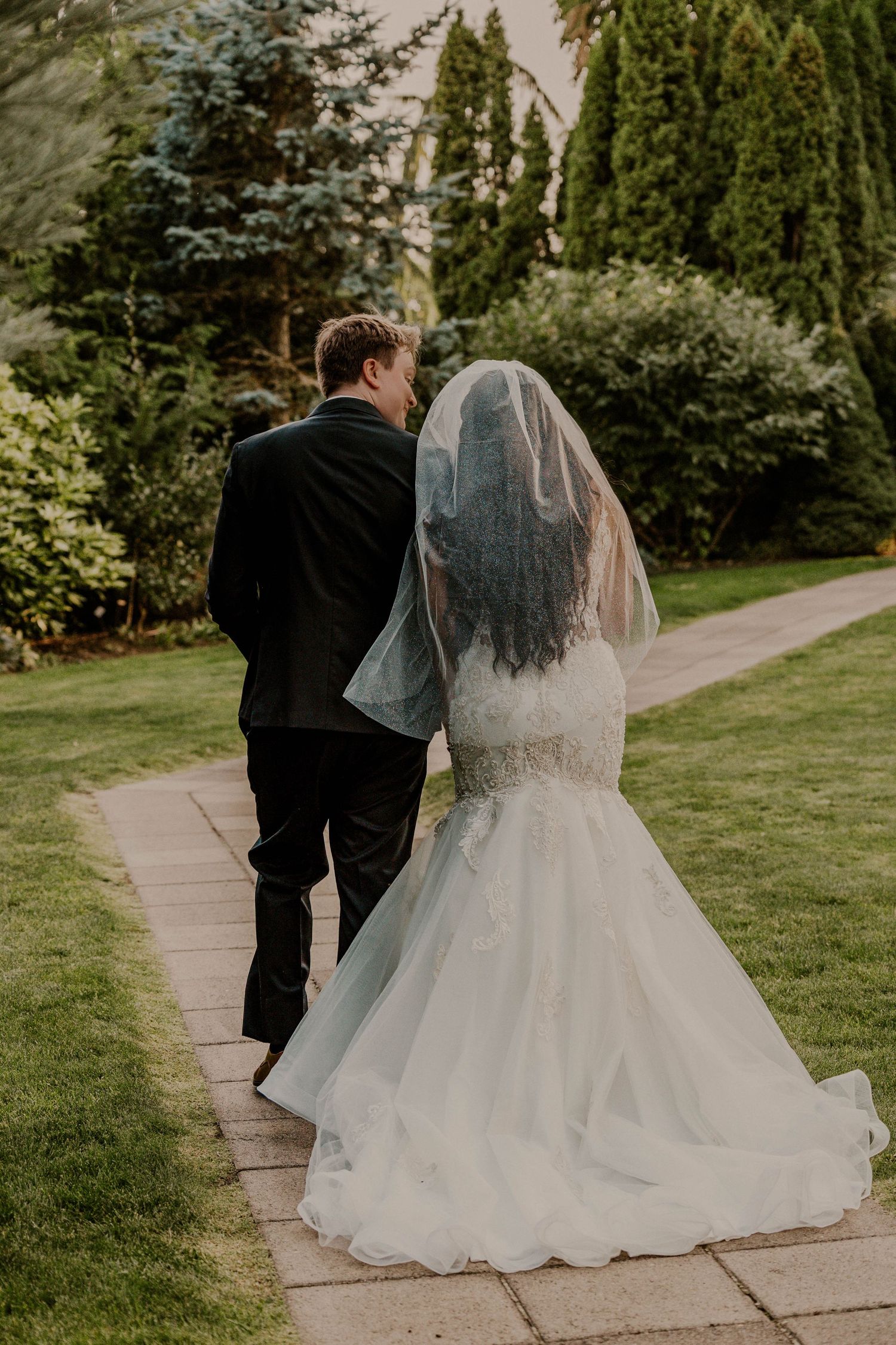 Bride and groom walking hand in hand along a garden path, bride wearing a lace mermaid gown with veil.
