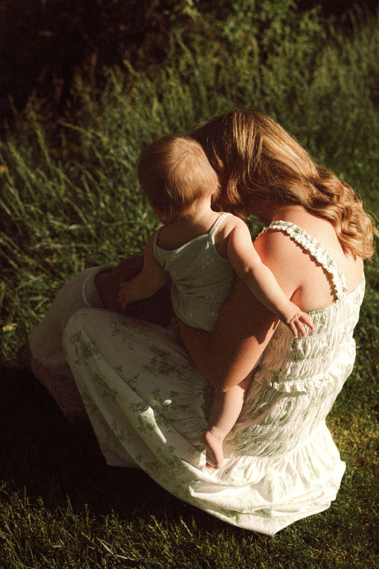 Mother in floral dress cradles baby on grass in warm sunlit outdoor setting.
