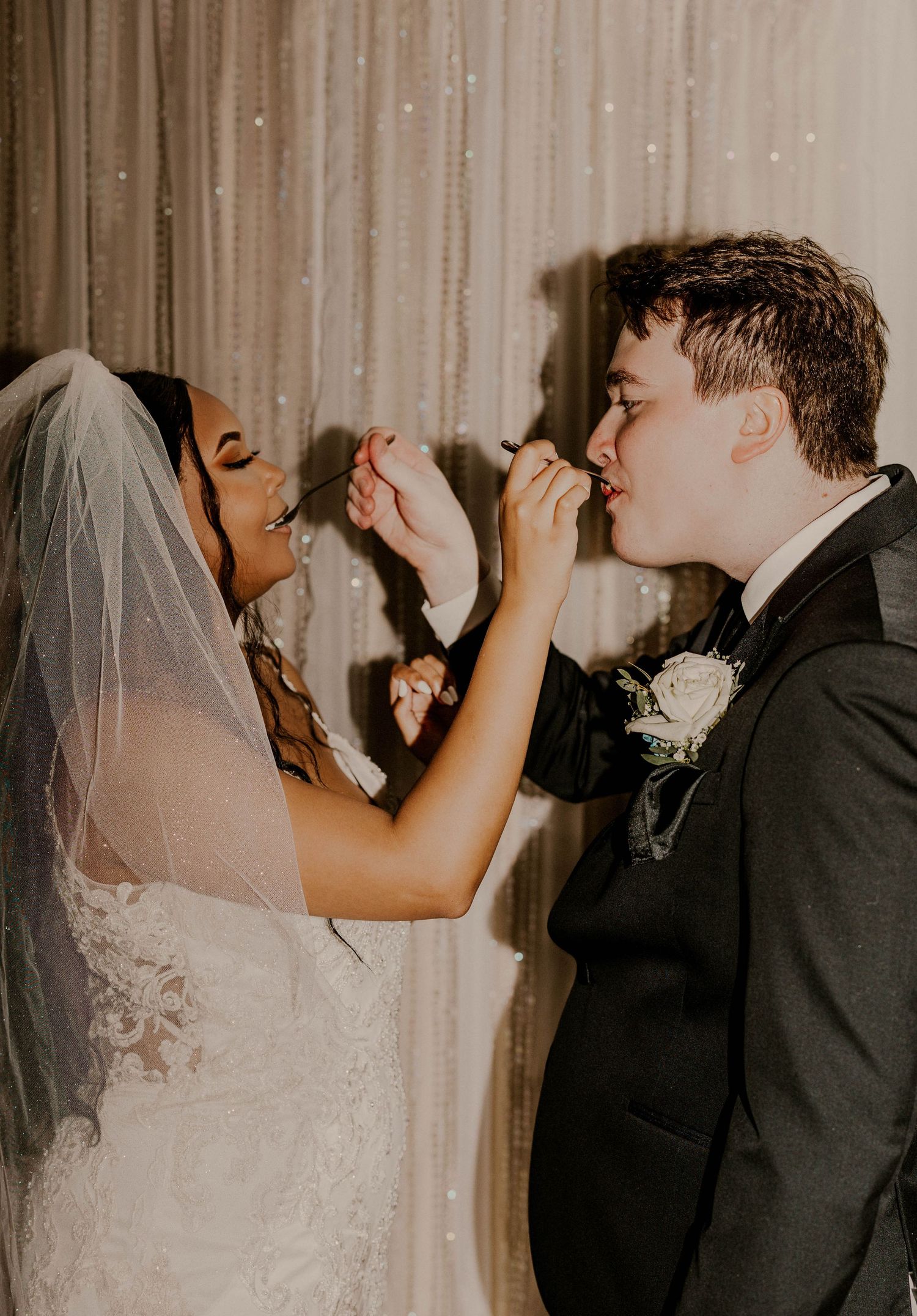 Bride feeding groom a piece of wedding cake at their reception in front of a shimmering backdrop.