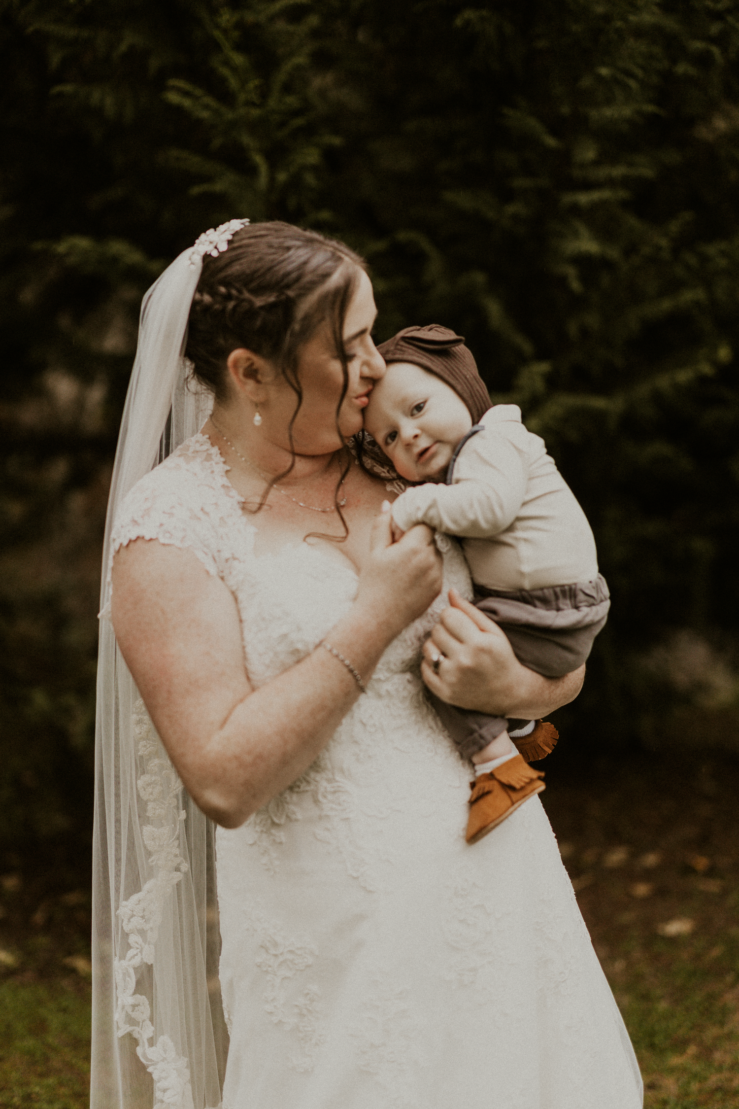 Bride in lace wedding gown tenderly holding and kissing a sleeping baby.