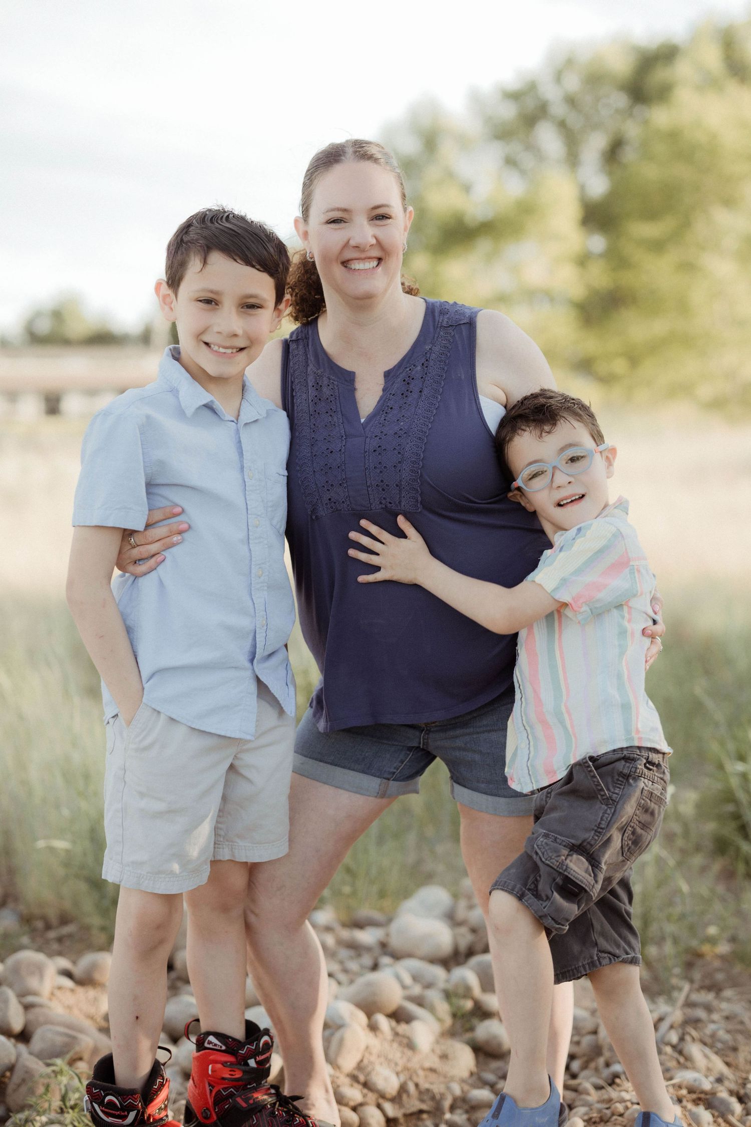 A smiling mother poses outdoors with her two young sons on a rocky natural landscape.