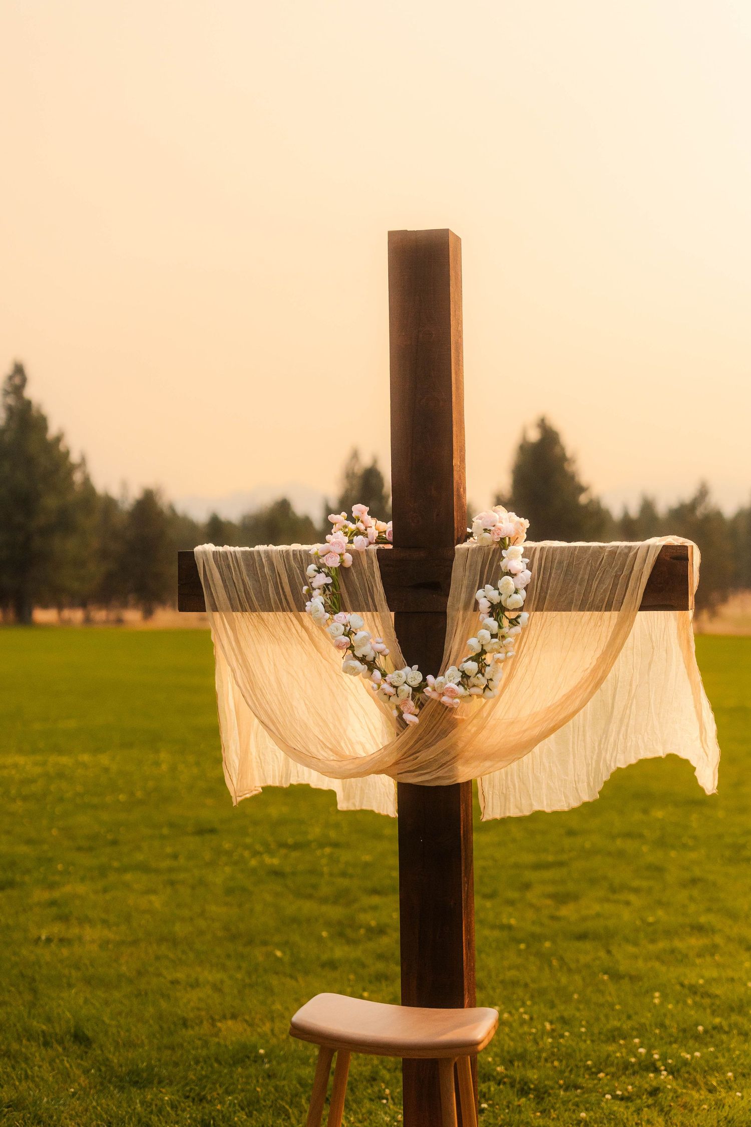 Wooden cross decorated with floral crown and draped fabric at an outdoor wedding ceremony venue.