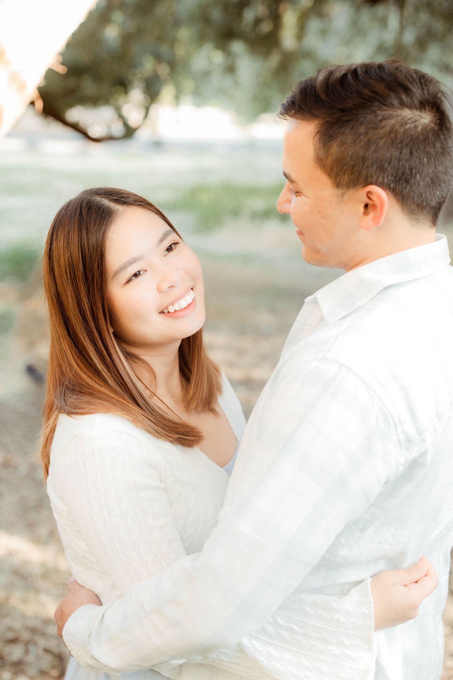 Smiling Asian woman embraces her partner outdoors during a romantic engagement photo session under a tree.