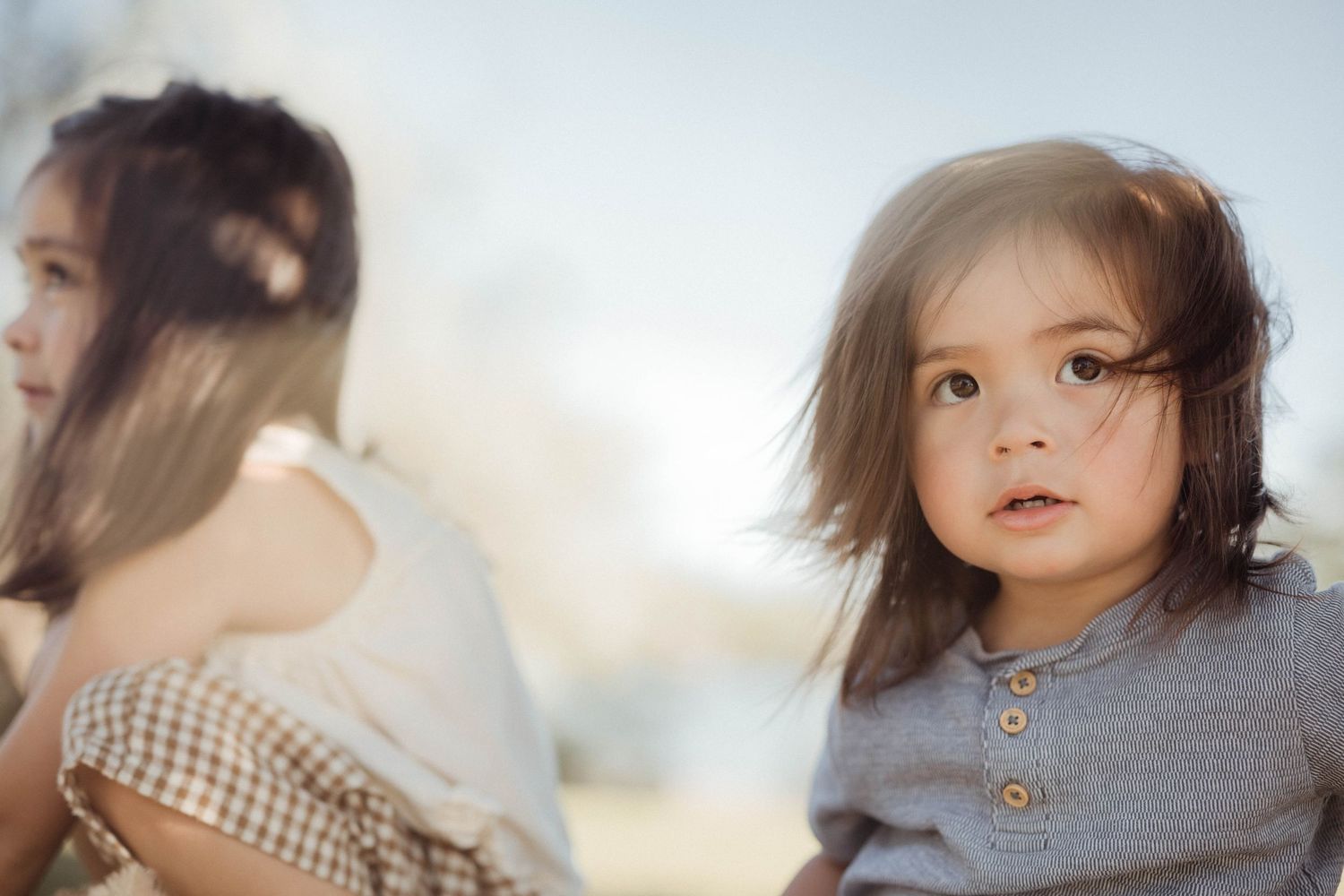 A young toddler with dark hair in a gray henley shirt looks at the camera outdoors in soft light.