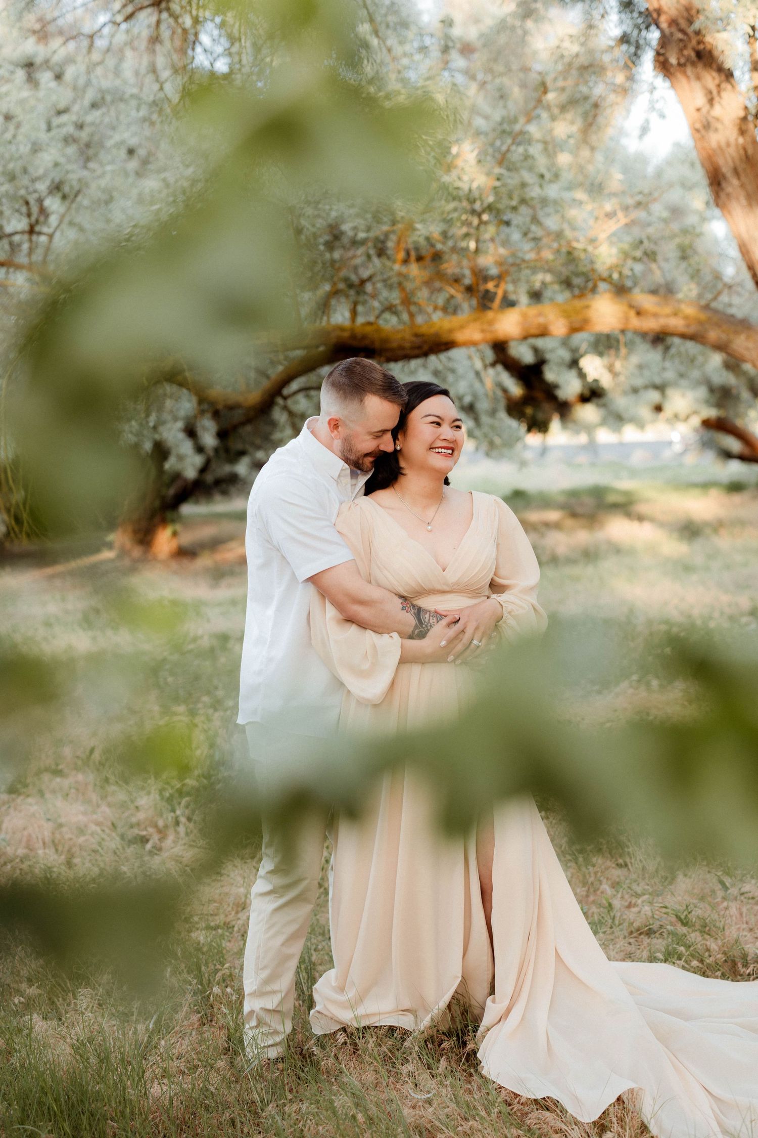 Couple embracing and laughing in golden hour light, framed by green leaves in a natural setting.