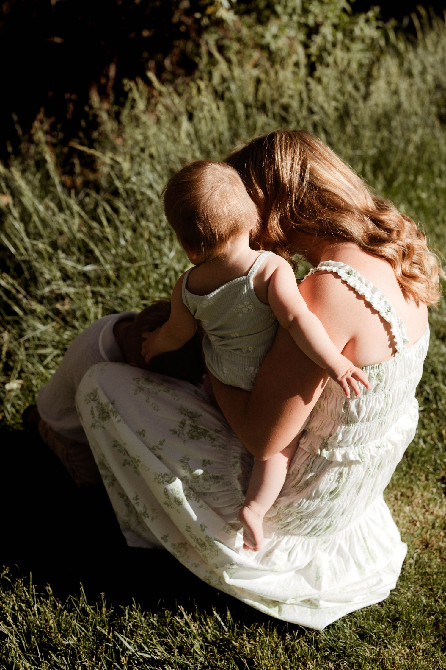A mother in a white floral dress sits on grass cuddling her baby in warm golden sunlight from behind.