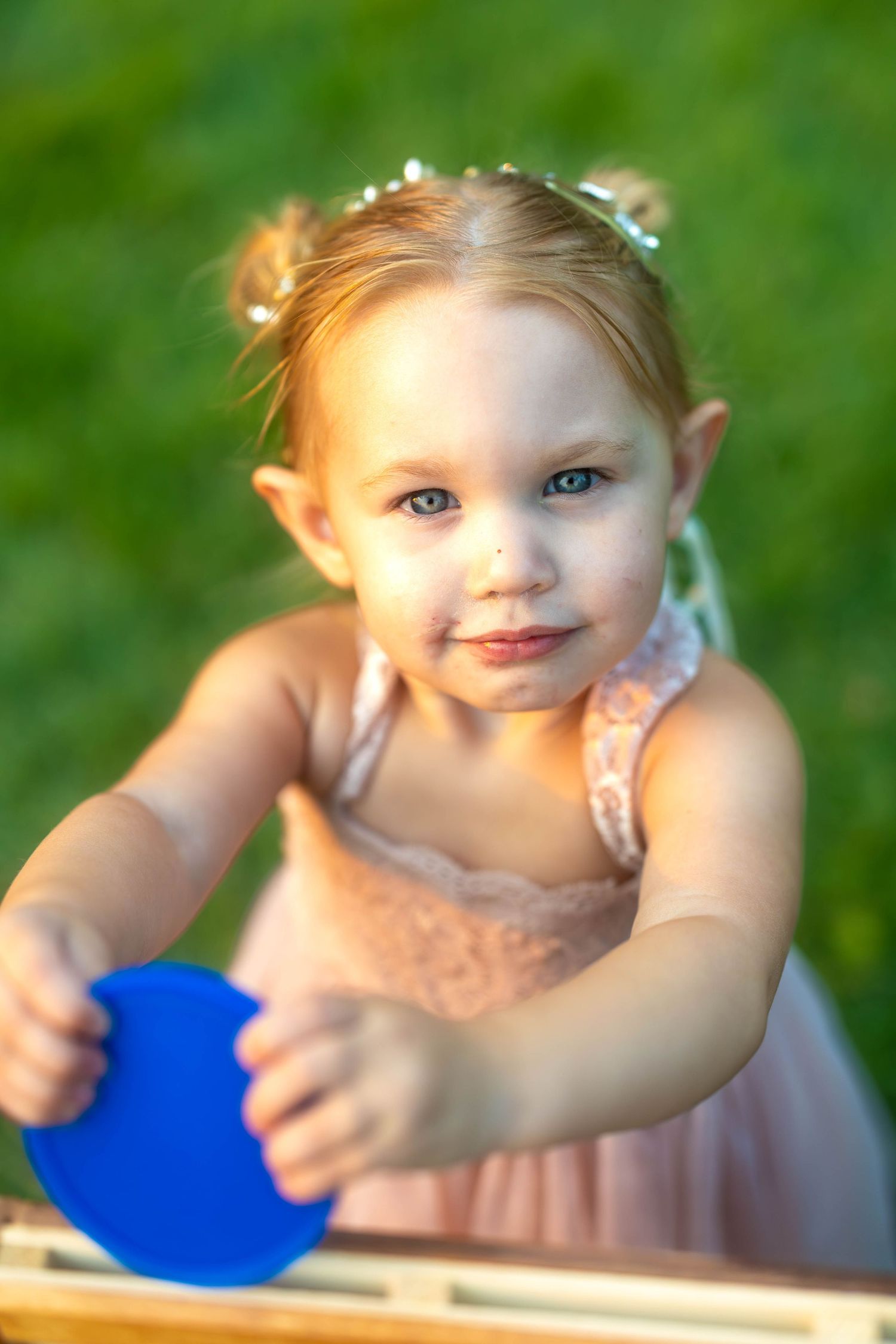 Adorable toddler girl in pink dress with floral crown holding a blue shape outdoors on green grass.