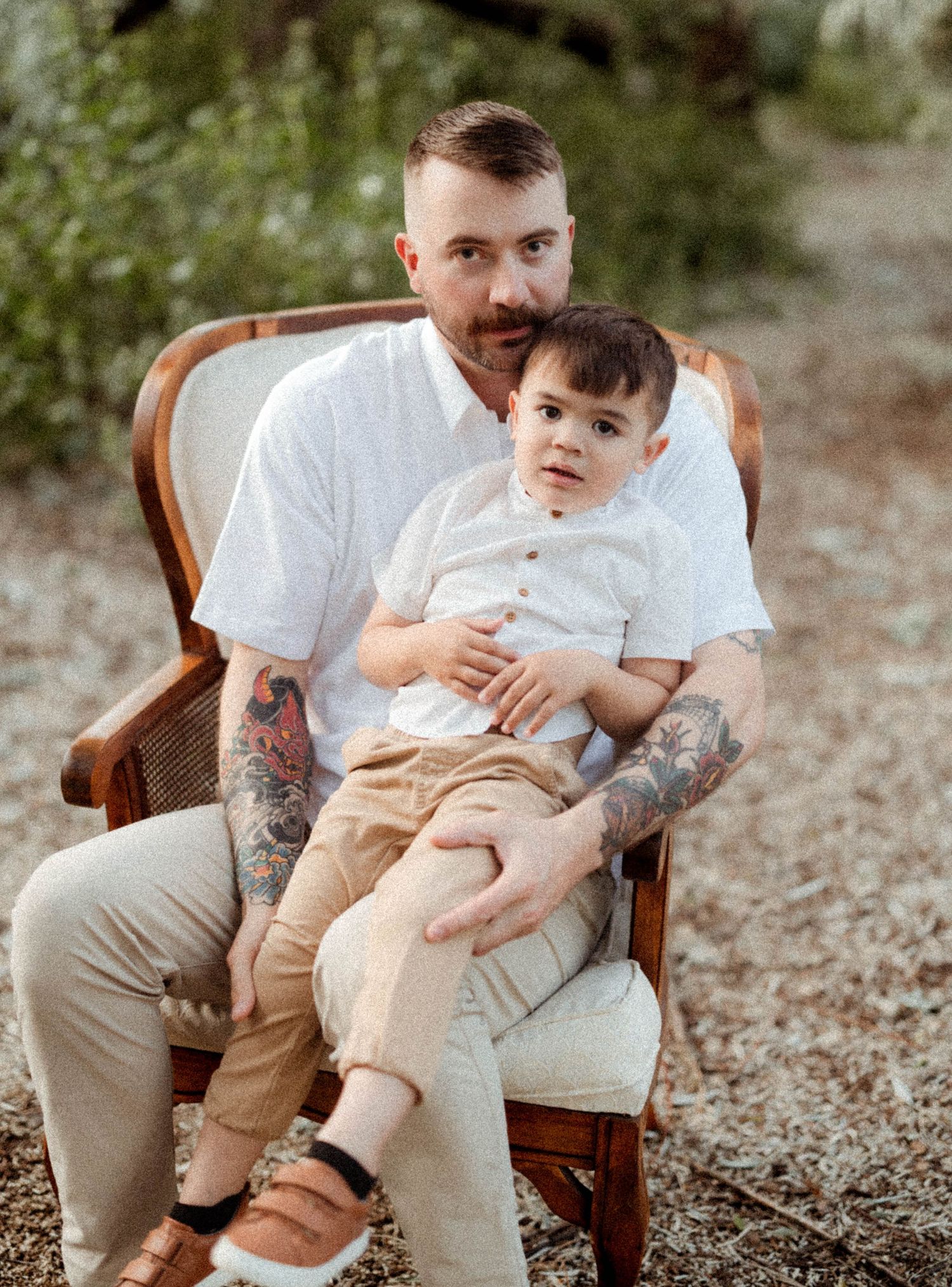 A tattooed father sits in a wooden chair holding his toddler son in an outdoor setting.