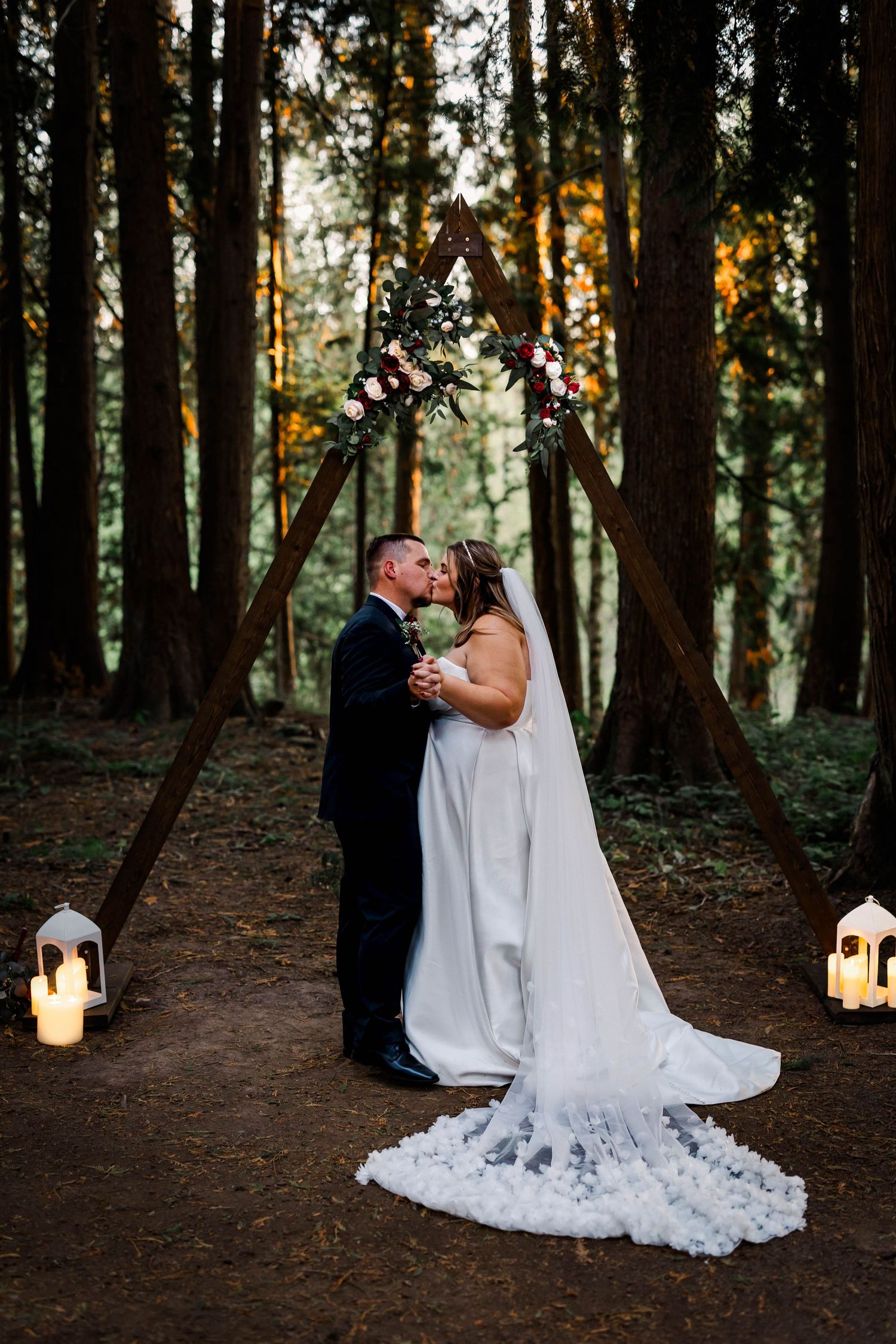 Bride and groom kissing under wooden triangular arch with flowers in forest.