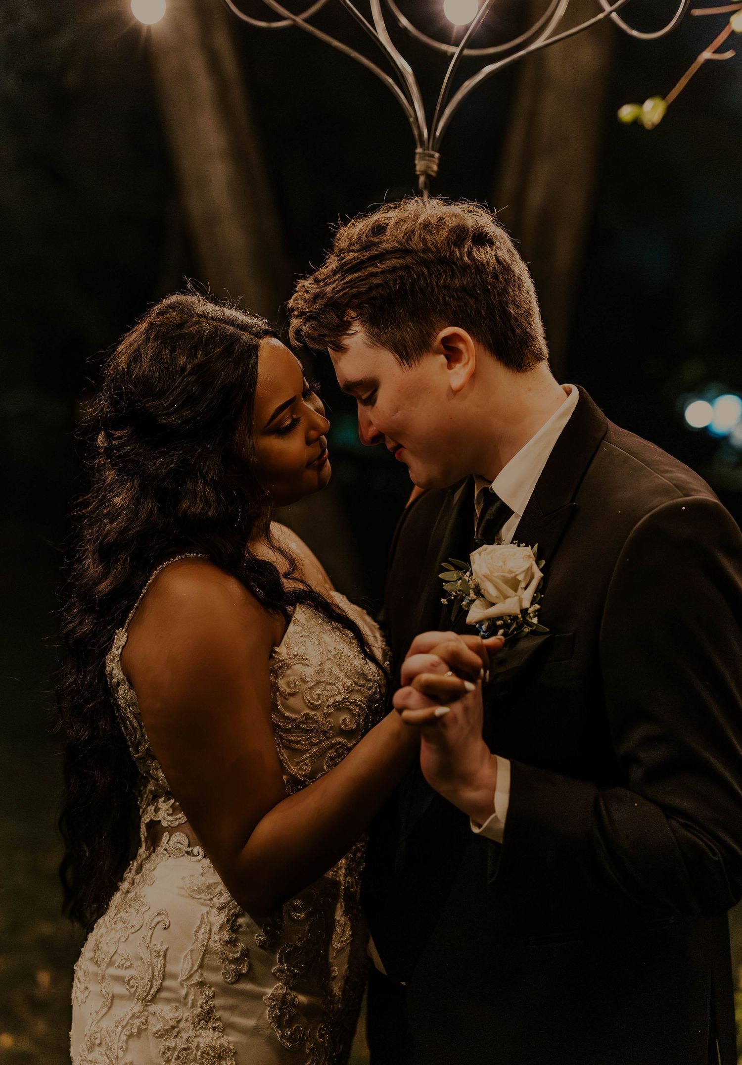 Newlyweds share a tender forehead touch while holding hands during their romantic nighttime wedding celebration.