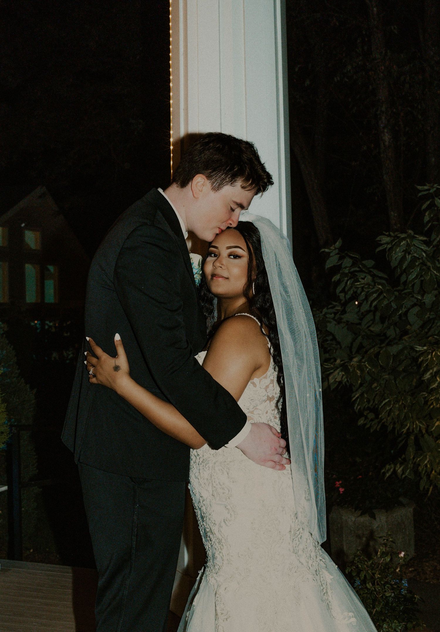 Bride and groom embracing outdoors at night, bride in lace mermaid gown with veil flowing.