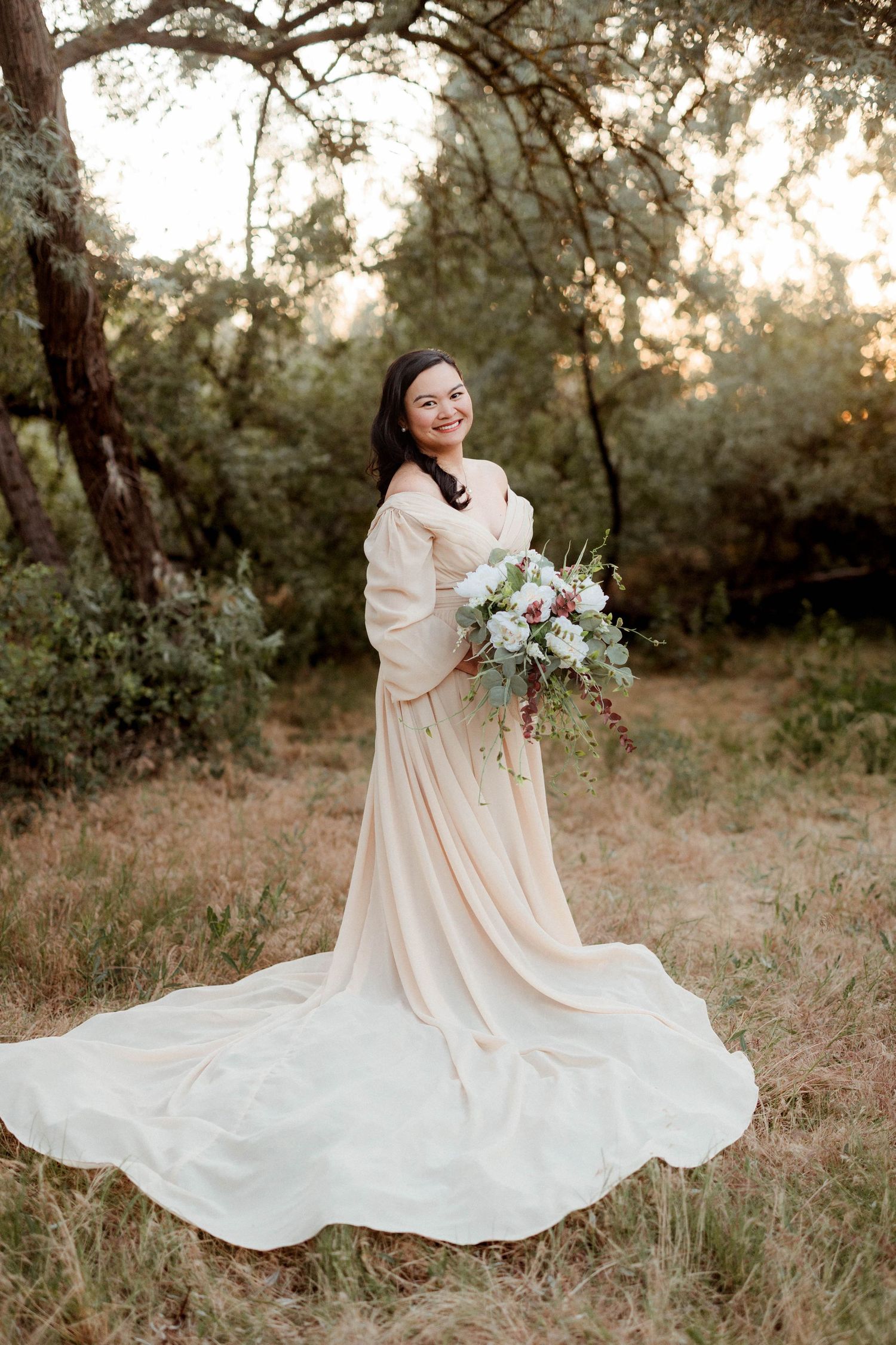 Smiling bride in off-shoulder champagne gown holding cascading bouquet in sun-dappled woodland setting.