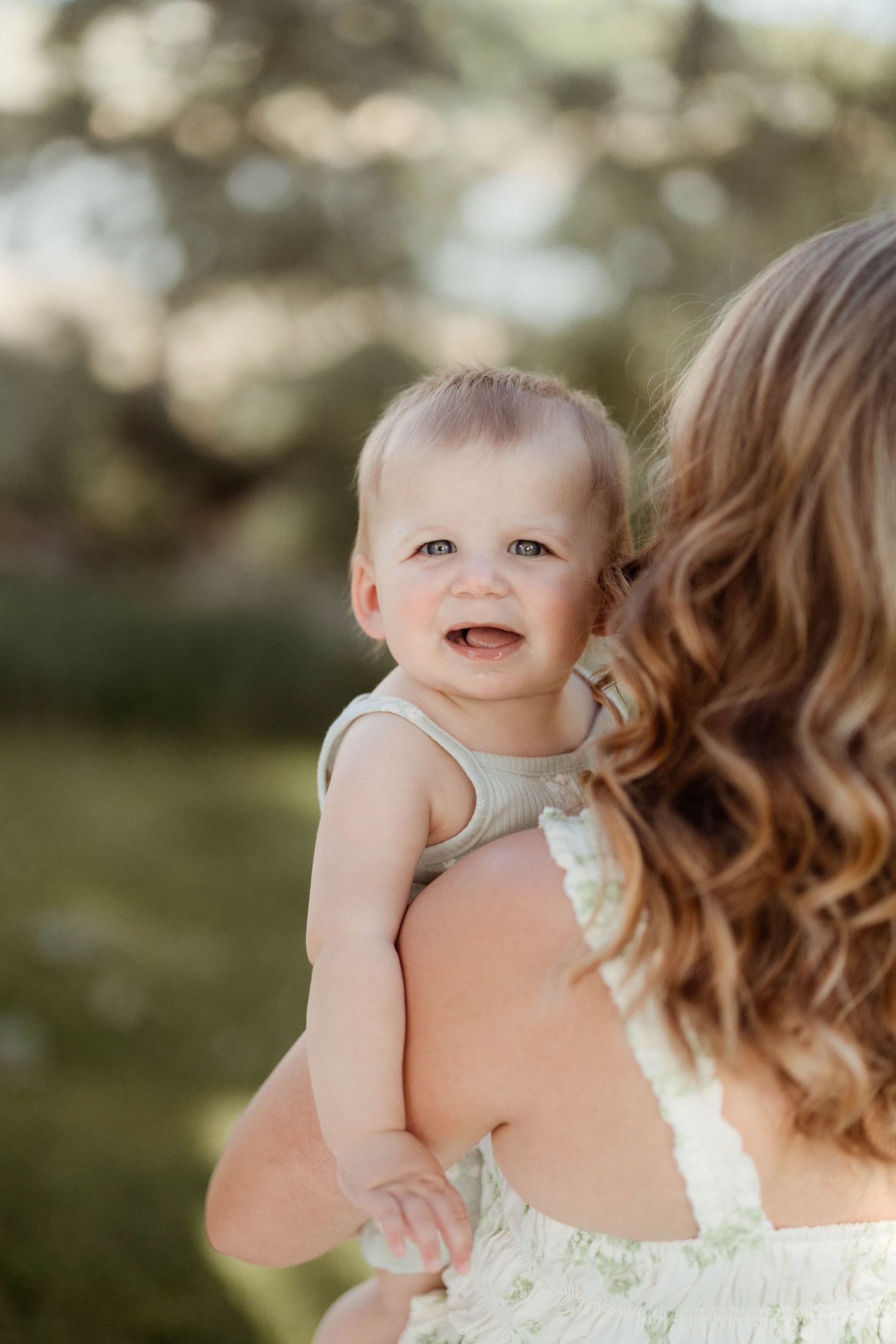 A smiling baby with blue eyes rests on a mothers shoulder in a sunny park.