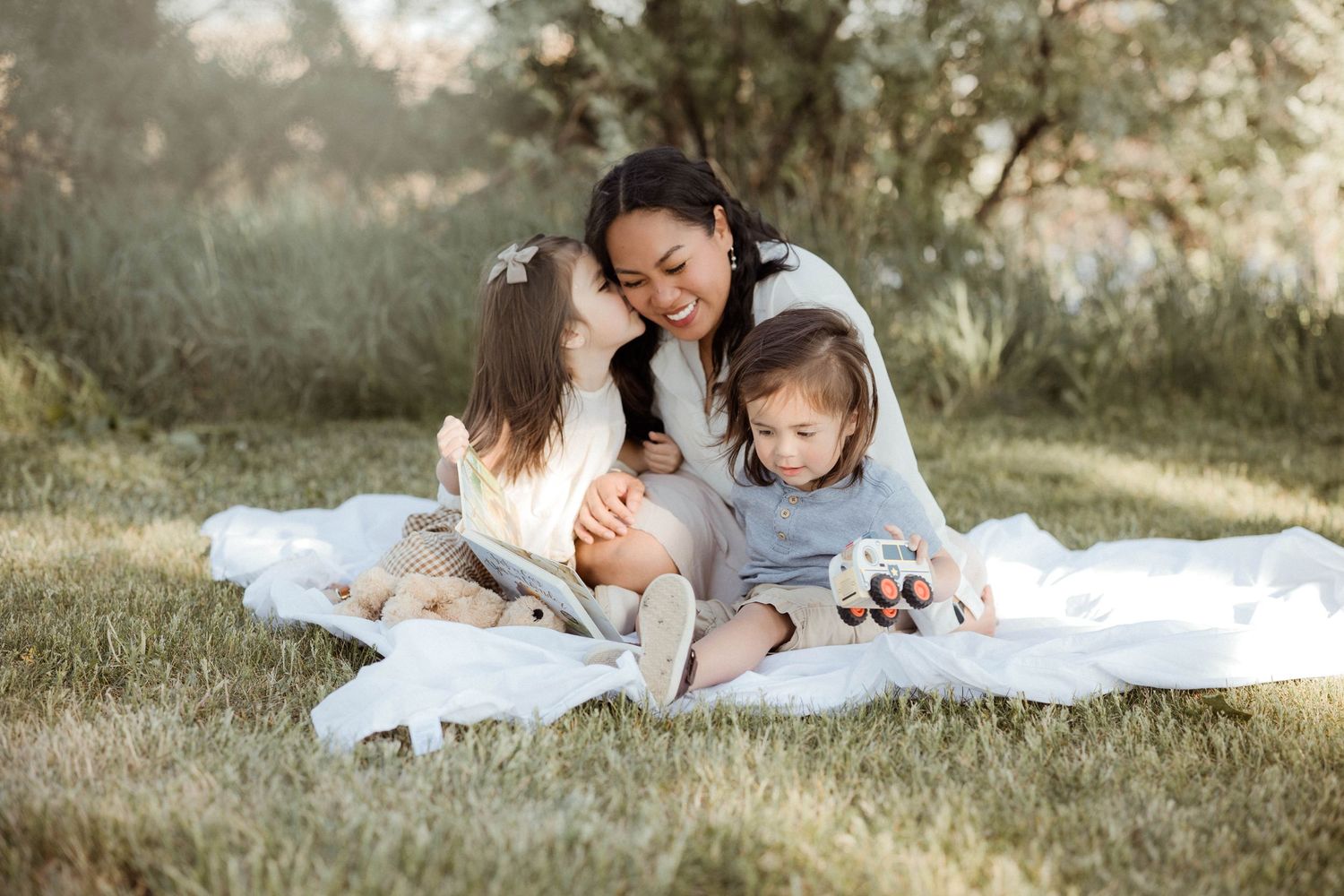 Mother sitting on blanket in park with two young children snuggled close in warm natural light.