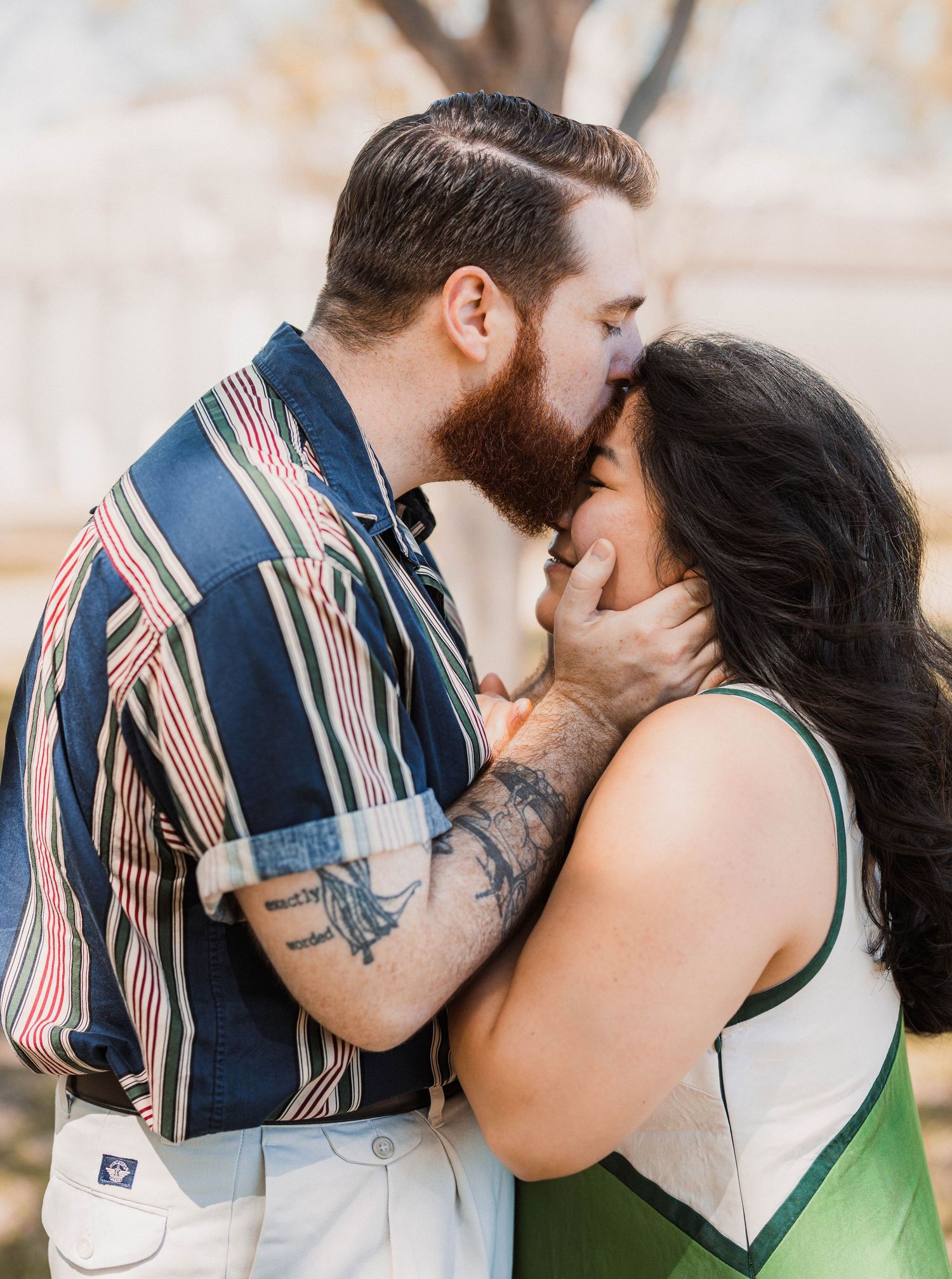 Bearded tattooed man tenderly kissing woman with dark hair outdoors in warm light.