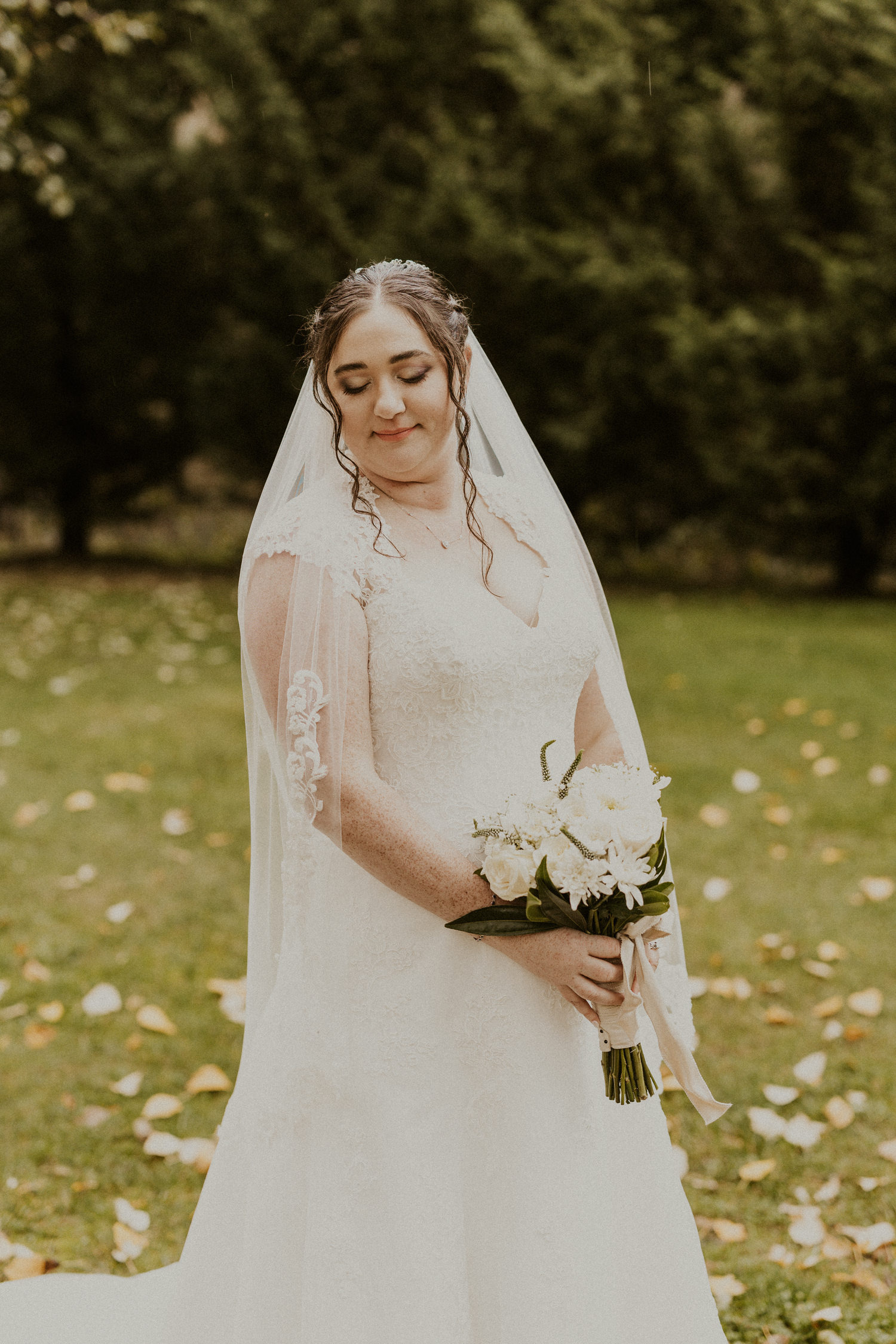 Bride in lace gown holding white bouquet with eyes closed in green garden.