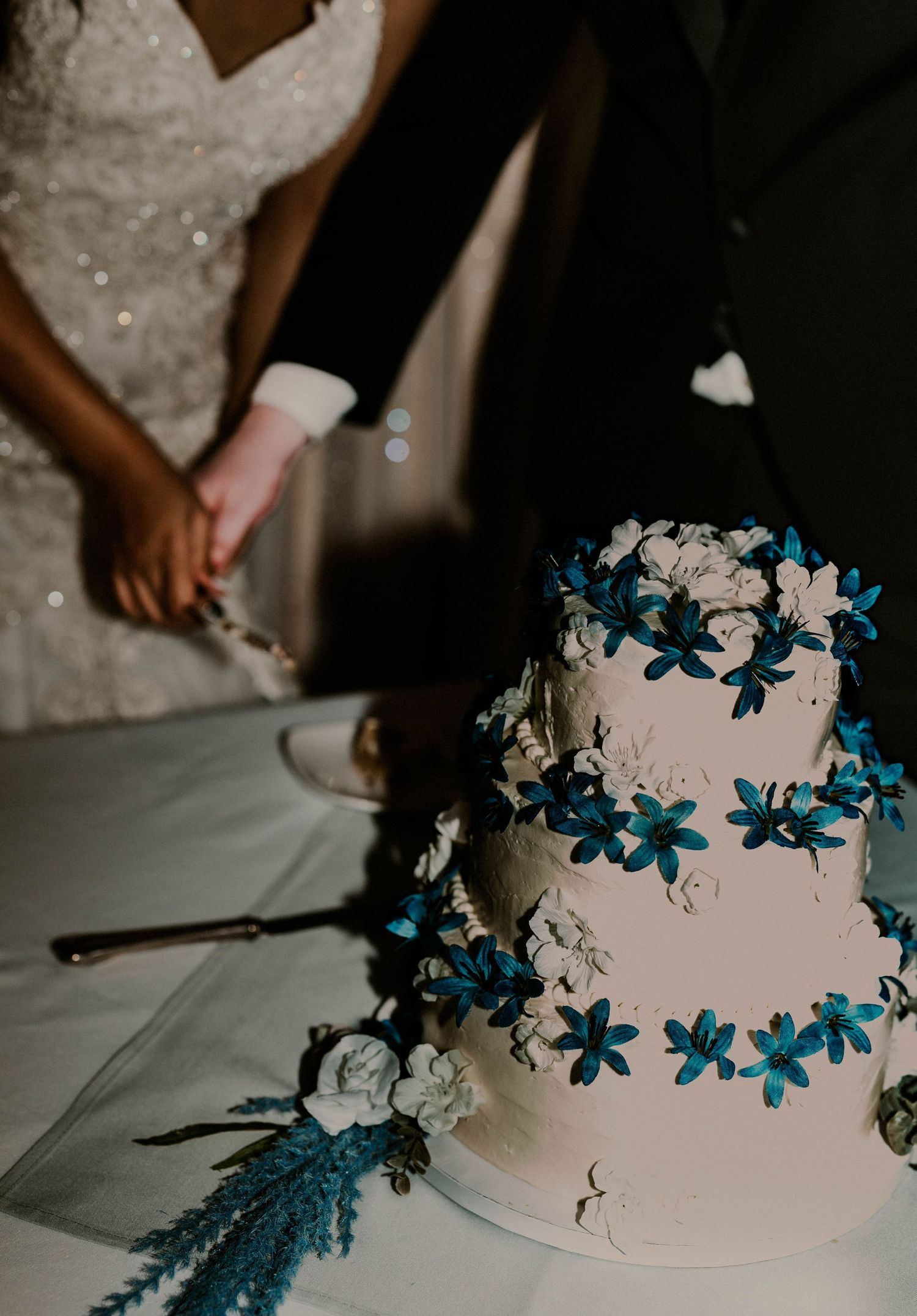 Two-tier white wedding cake decorated with blue and white flowers as couple prepares to cut it.
