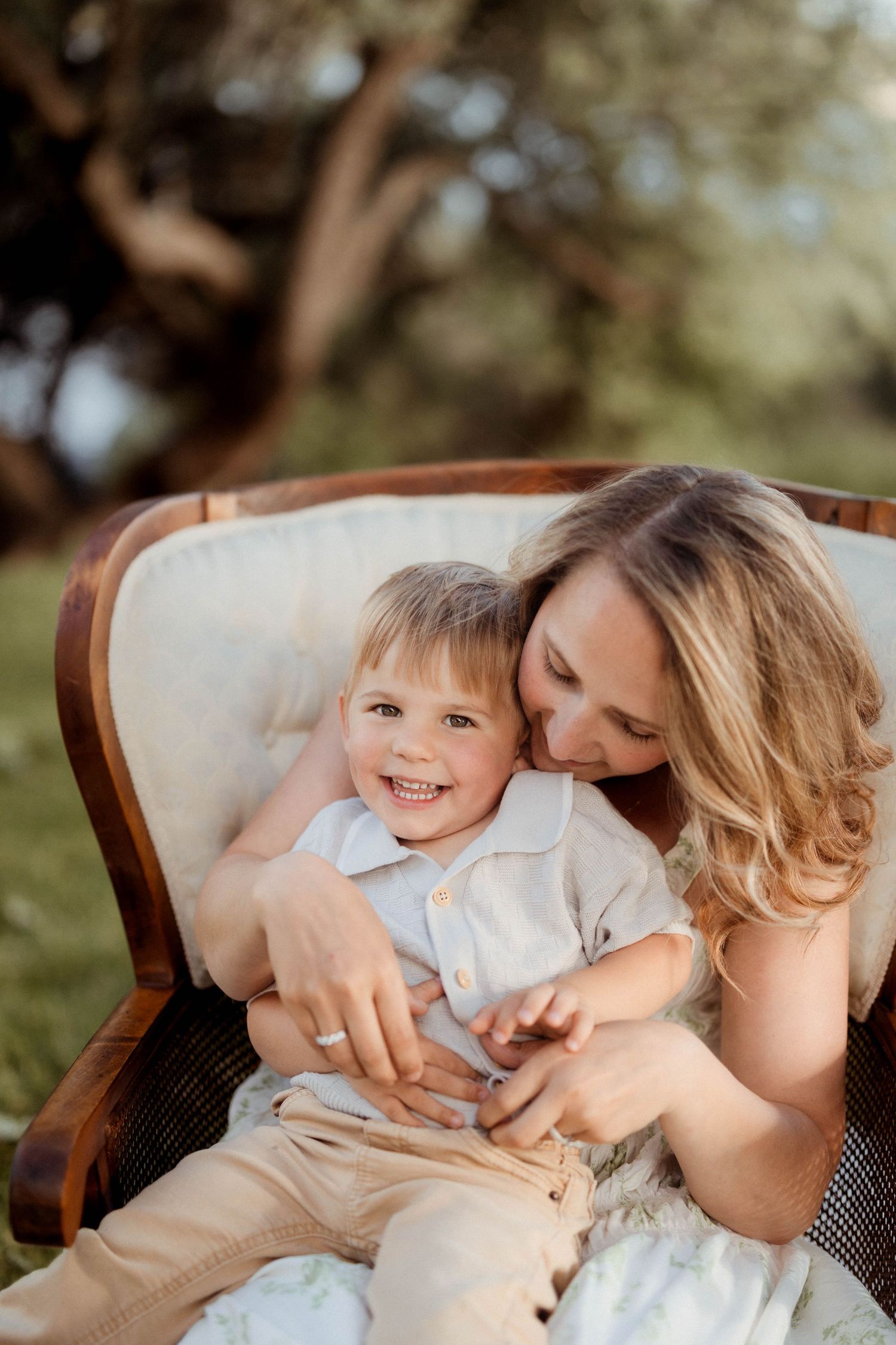 Laughing boy sits on mothers lap in vintage chair during warm outdoor family portrait.