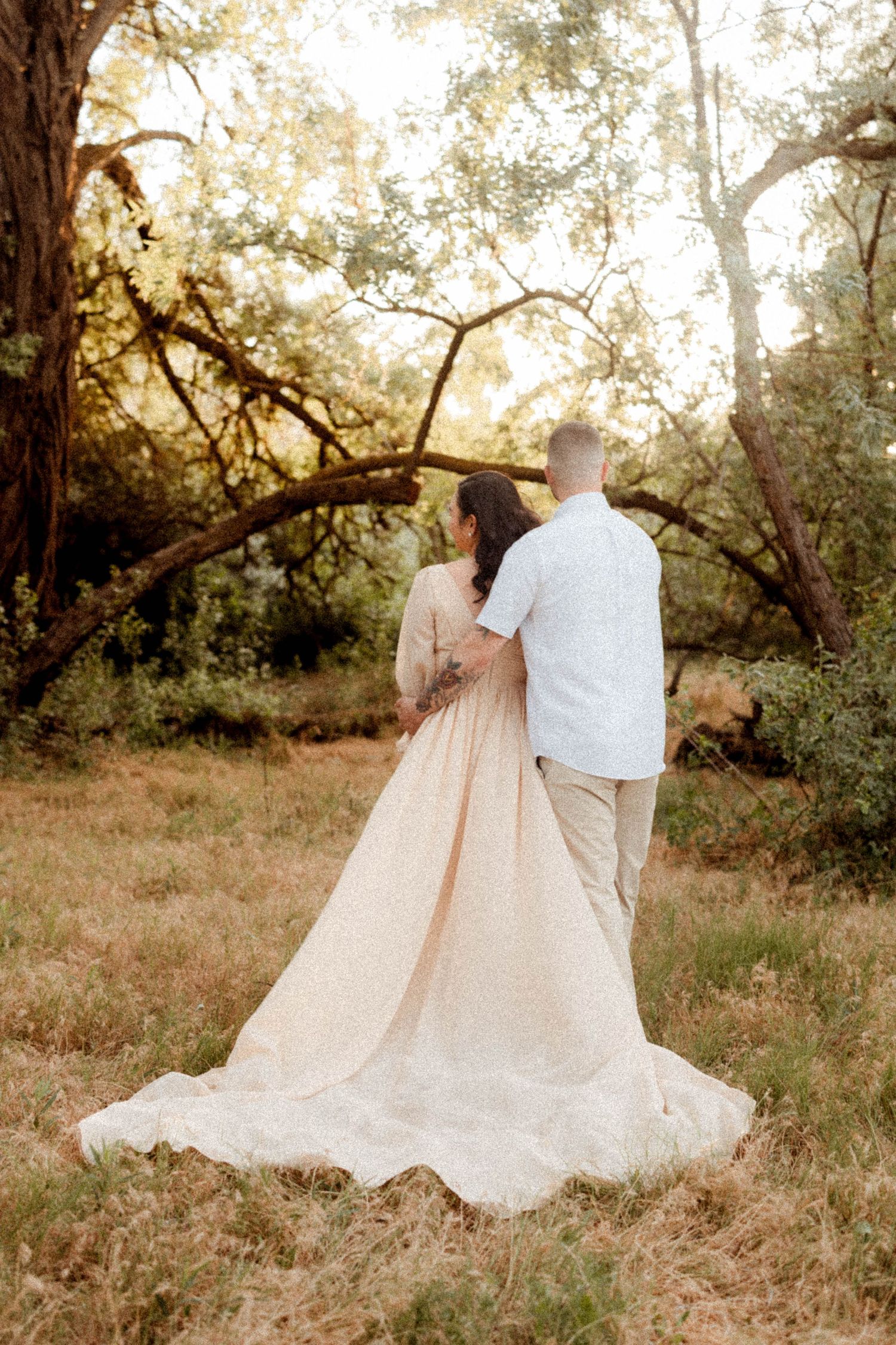 Bride in a flowing champagne gown and groom walking hand in hand through a sunlit forest.