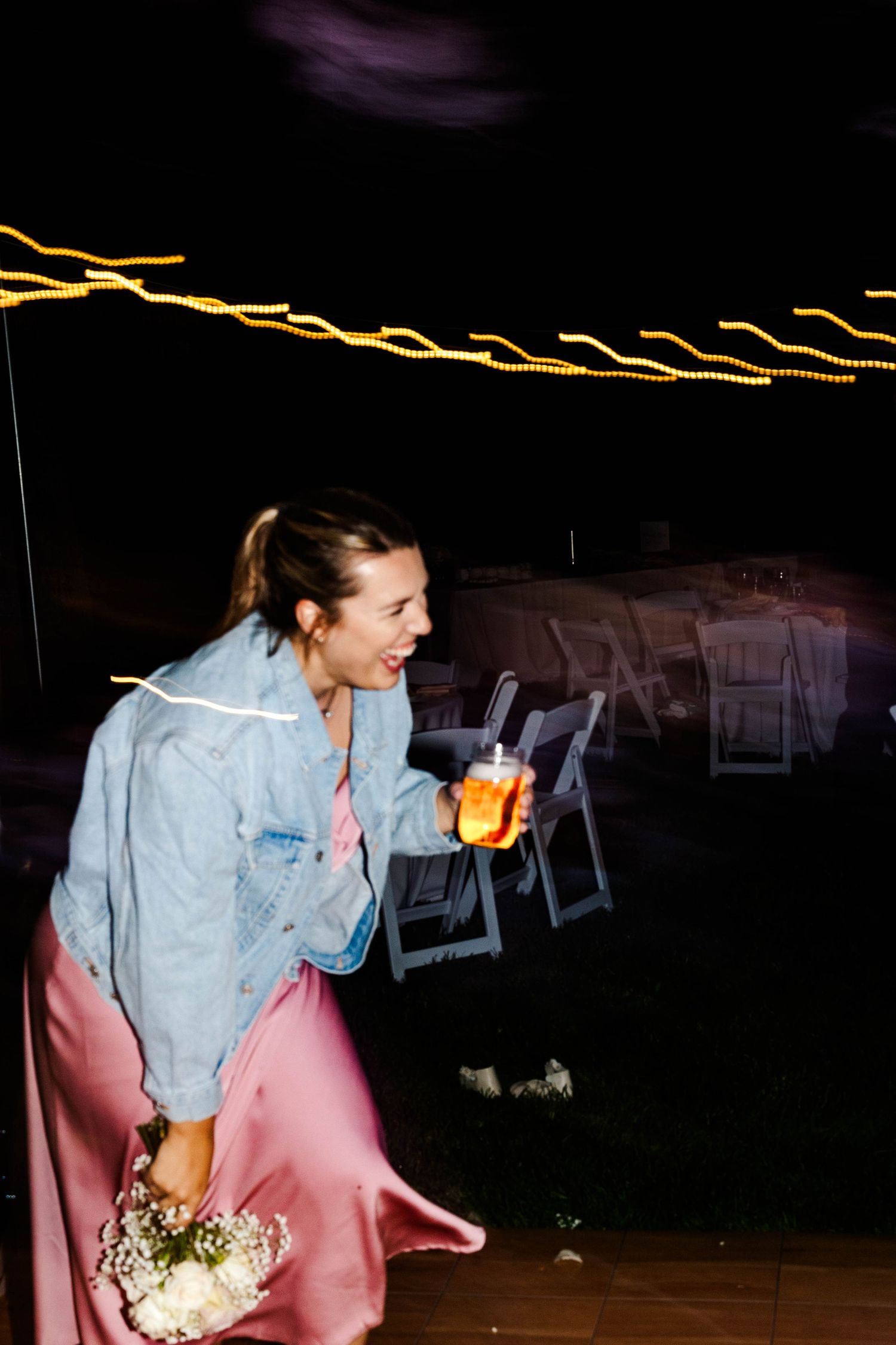 Joyful bridesmaid in pink satin skirt and denim jacket holding bouquet and drink at outdoor wedding.