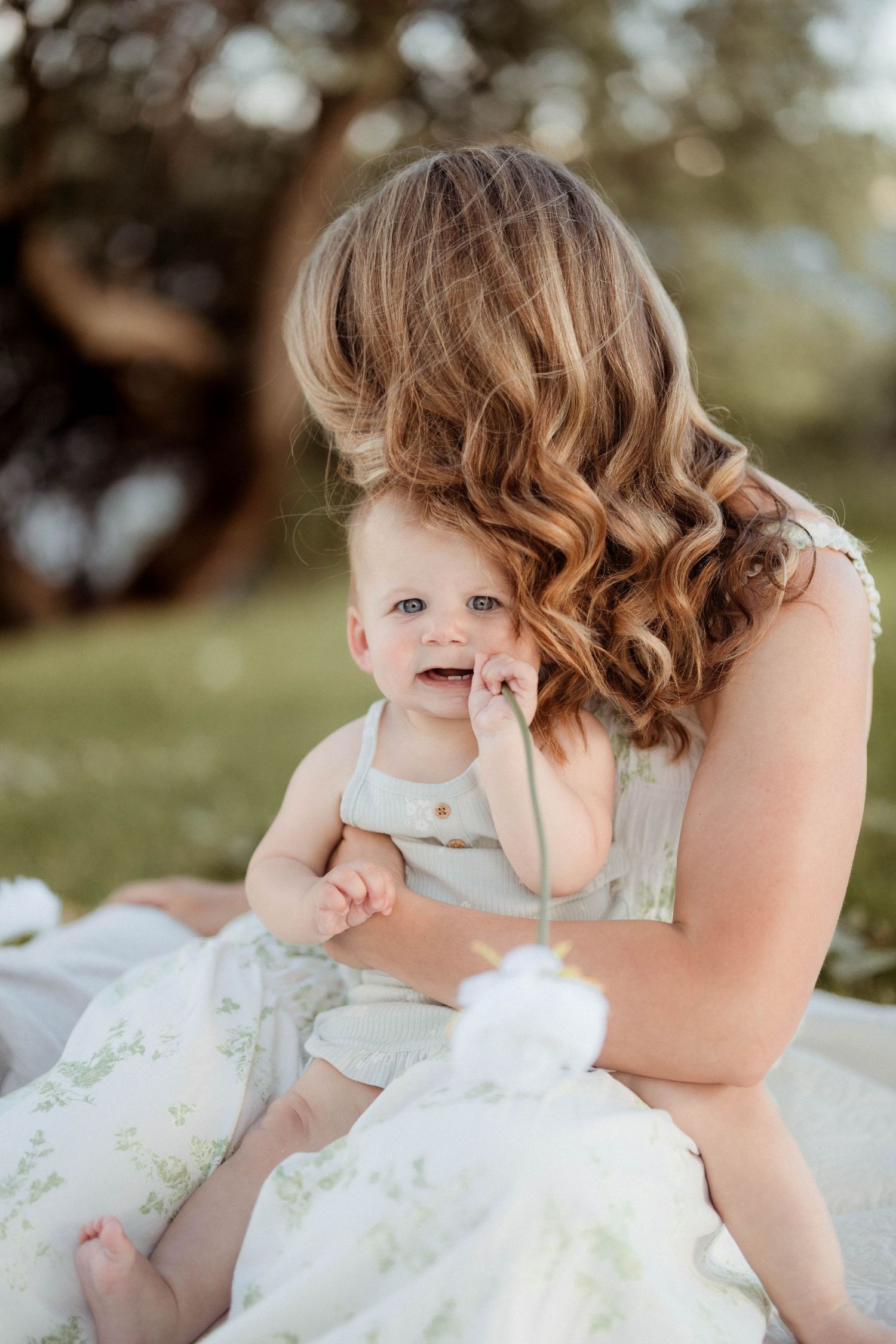 Mother with curly hair holds smiling baby in green floral outfit during golden hour outdoor photo session.