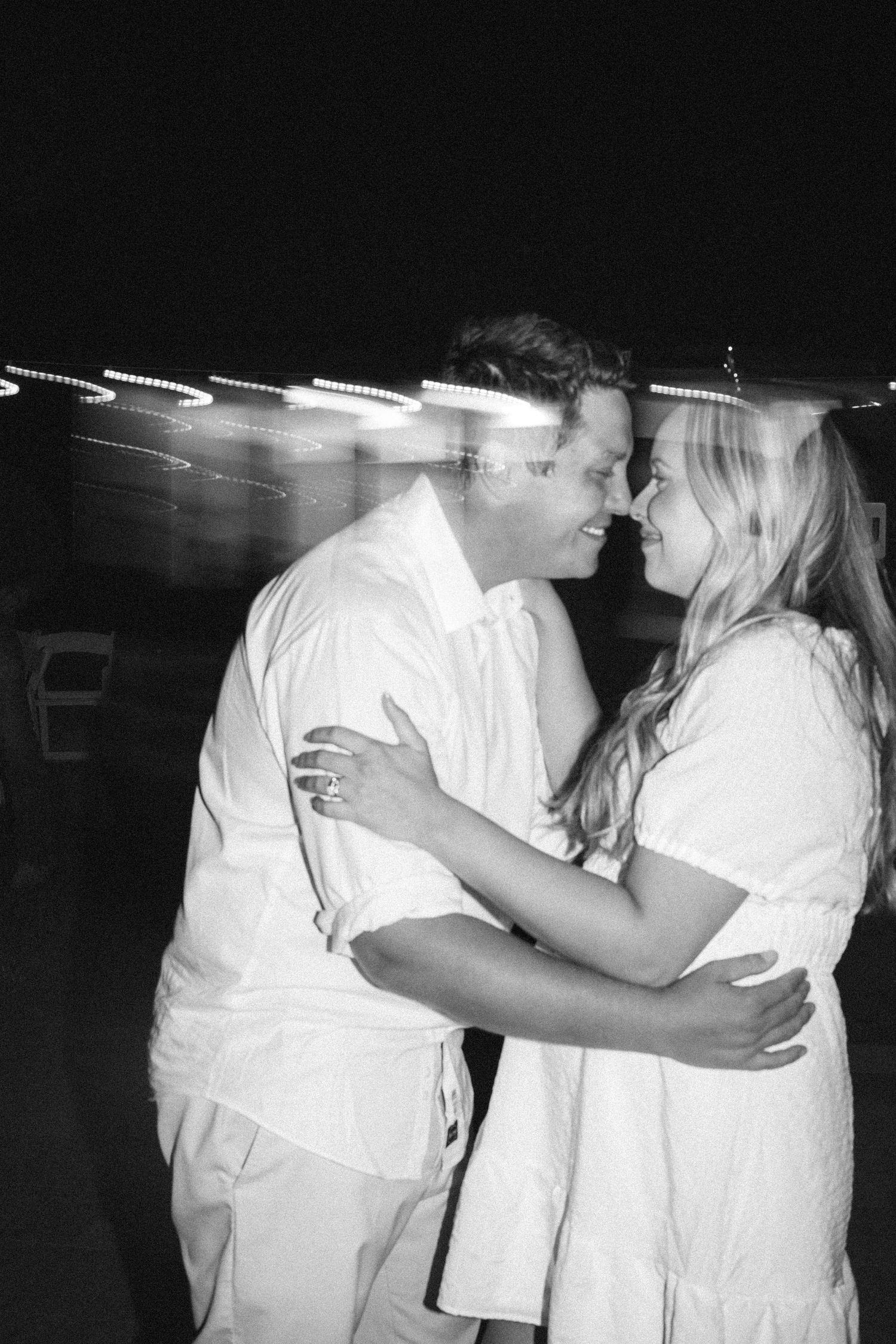 Black and white romantic photo of newlywed couple embracing and smiling under string lights at night.